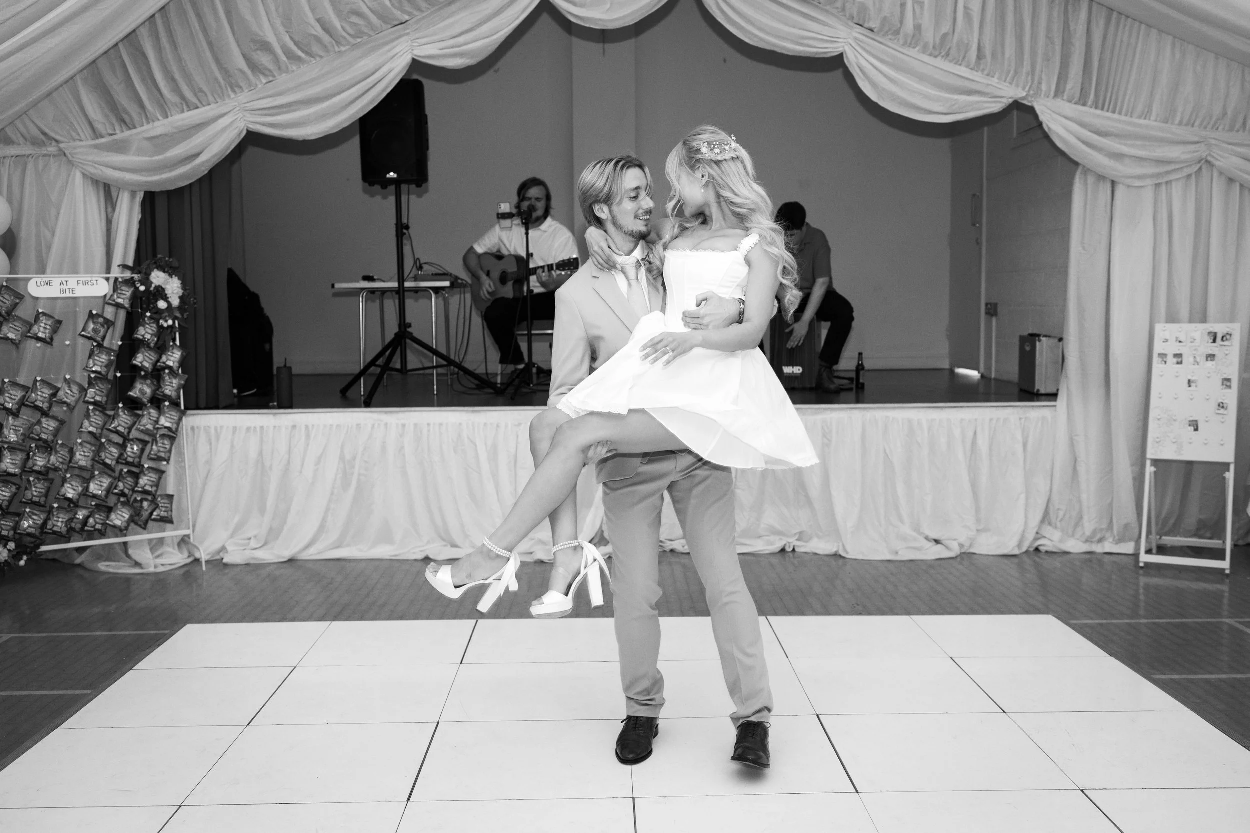 A man in a suit lifting a woman in a white dress on a dance floor, with a band playing instruments in the background at a wedding reception.