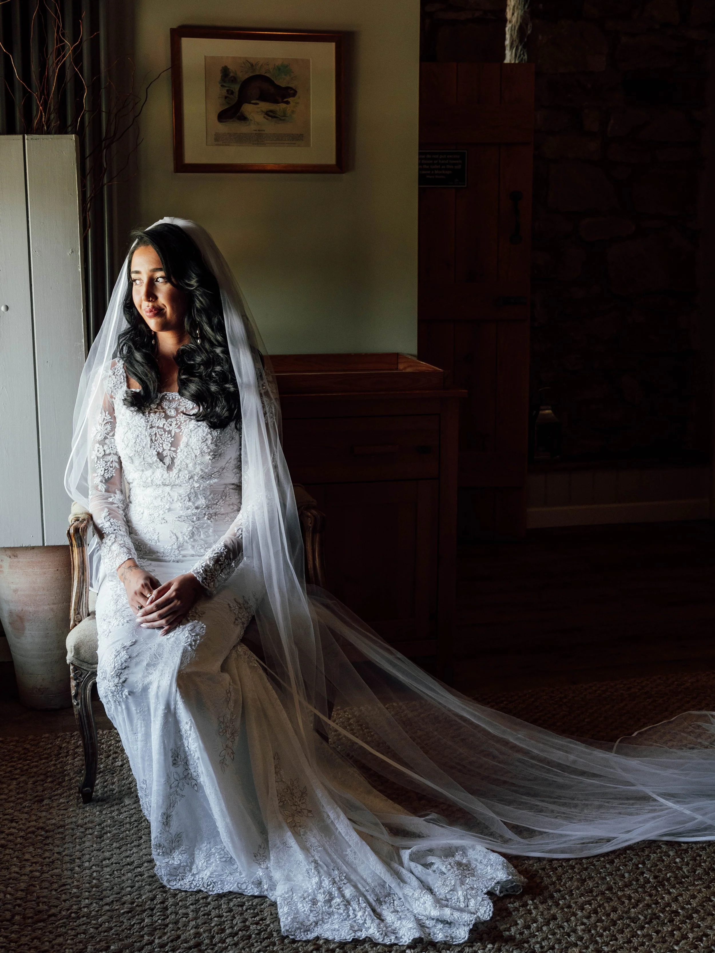 A bride in a lace wedding gown with long sleeves sits on a vintage chair, wearing a veil, inside a room with natural light coming through a window, surrounded by wooden furniture and a framed animal illustration on the wall.