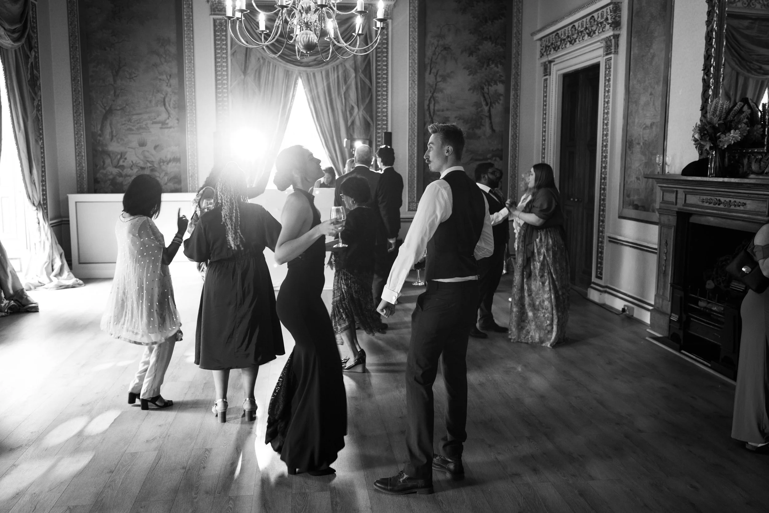People dancing and socializing at an elegant indoor event, with large windows, drapes, and ornate walls, captured in black and white.