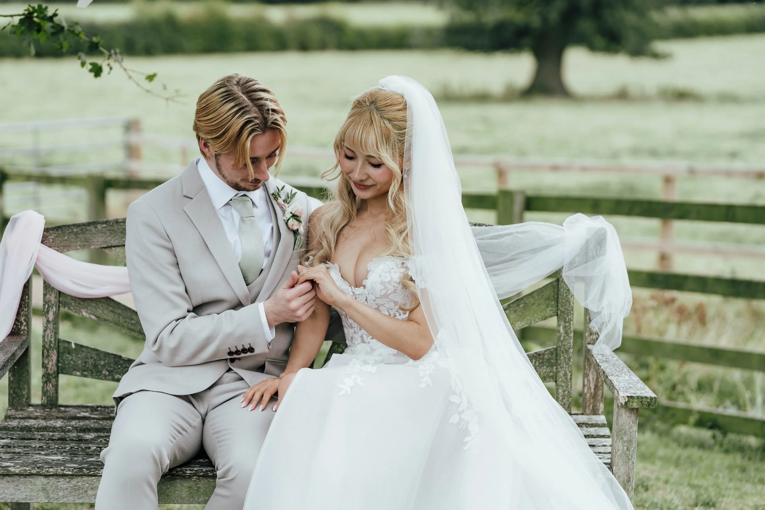 A bride and groom sitting on a rustic wooden bench outdoors, with the groom pinning a boutonniere on the bride's dress. The bride is wearing a white wedding gown and veil, and the groom is in a light gray suit. Green trees and a fence are in the back