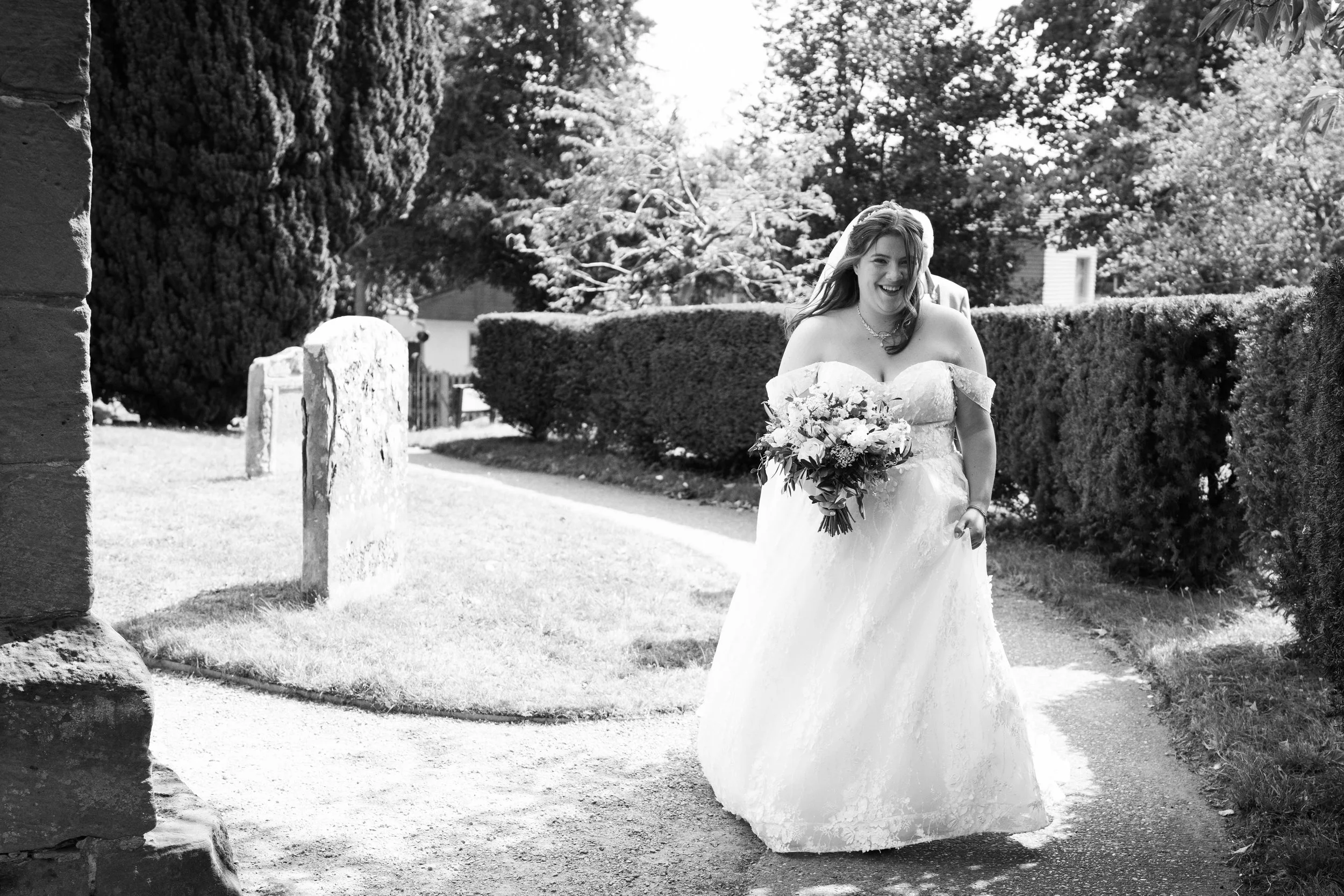 A joyful bride in a wedding dress walking outdoors with a bouquet of flowers, smiling and holding her dress.