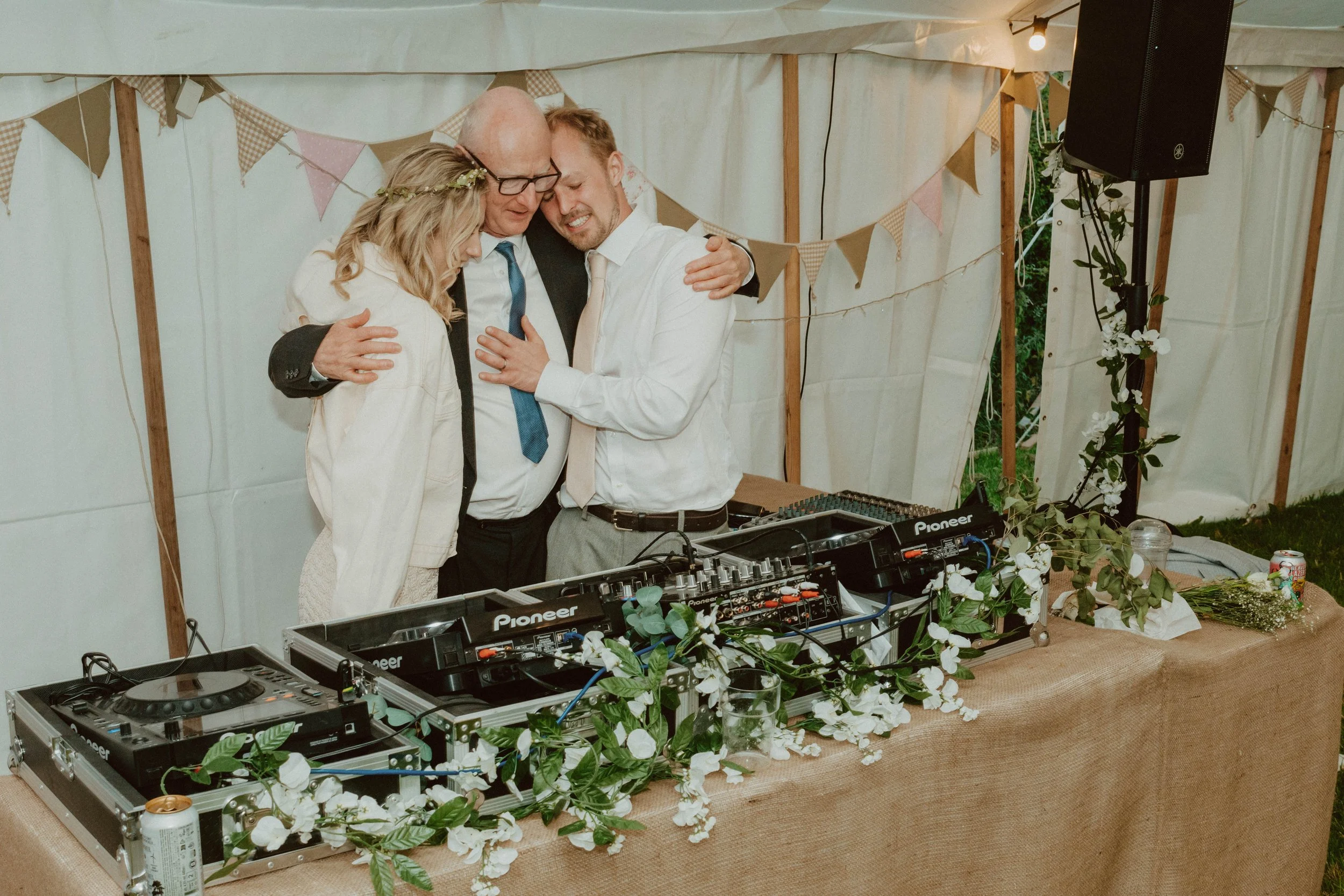 Three people, two men and a woman, hugging and holding hands in front of a DJ setup with DJ equipment and floral decorations inside a tent at a celebration or wedding.