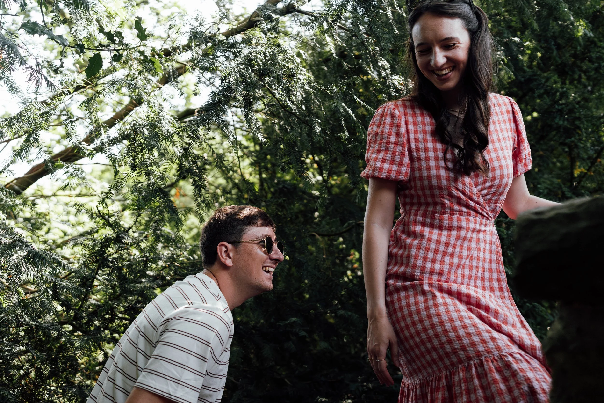 A young man wearing sunglasses and a striped shirt is kneeling and smiling at a young woman in a red gingham dress, who is standing outdoors amid greenery, both appearing happy and engaged.