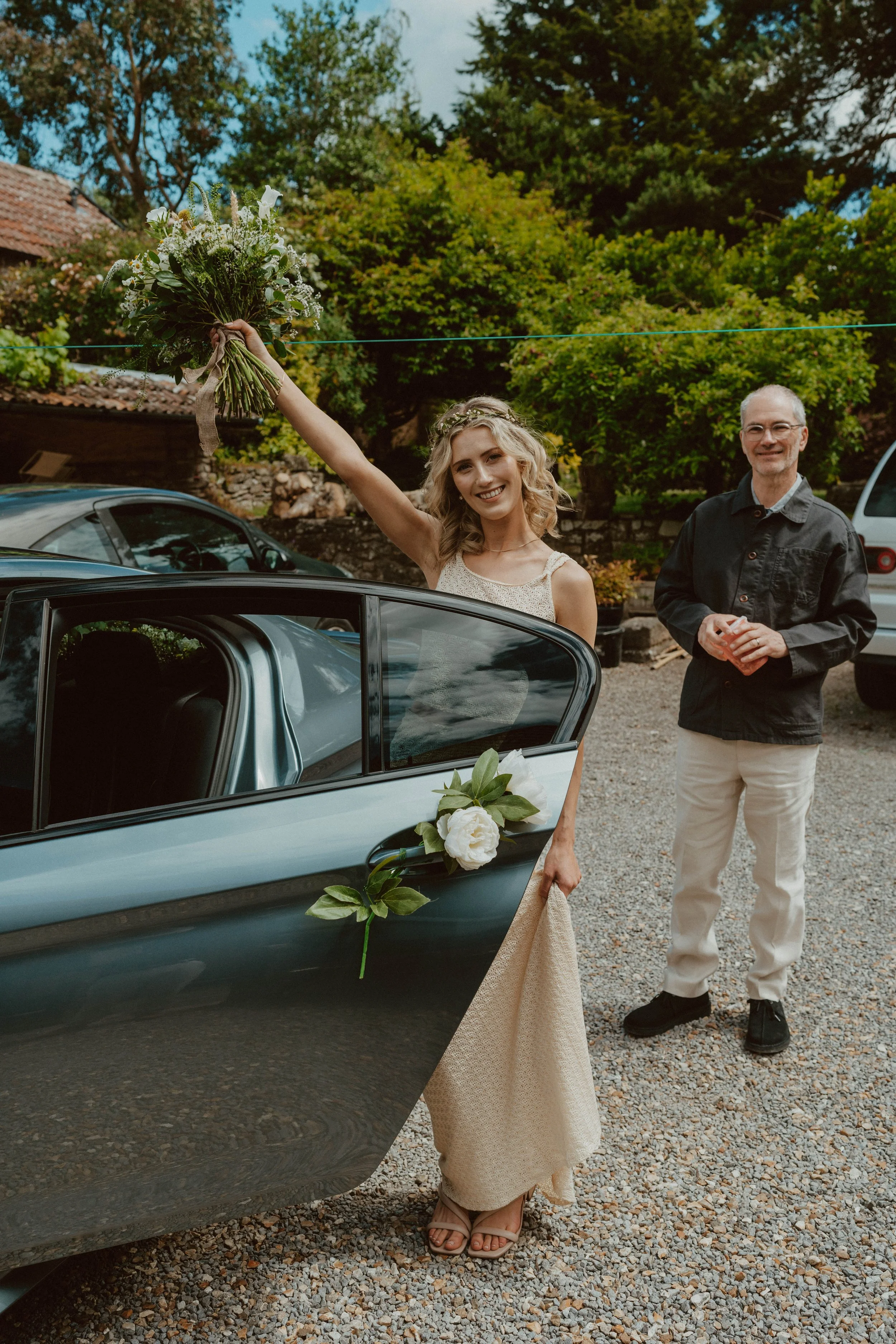 A smiling woman in a light-colored dress holding a bouquet of flowers raises her arm in celebration next to a decorated car, with a man standing nearby smiling, in an outdoor area with trees and old buildings.