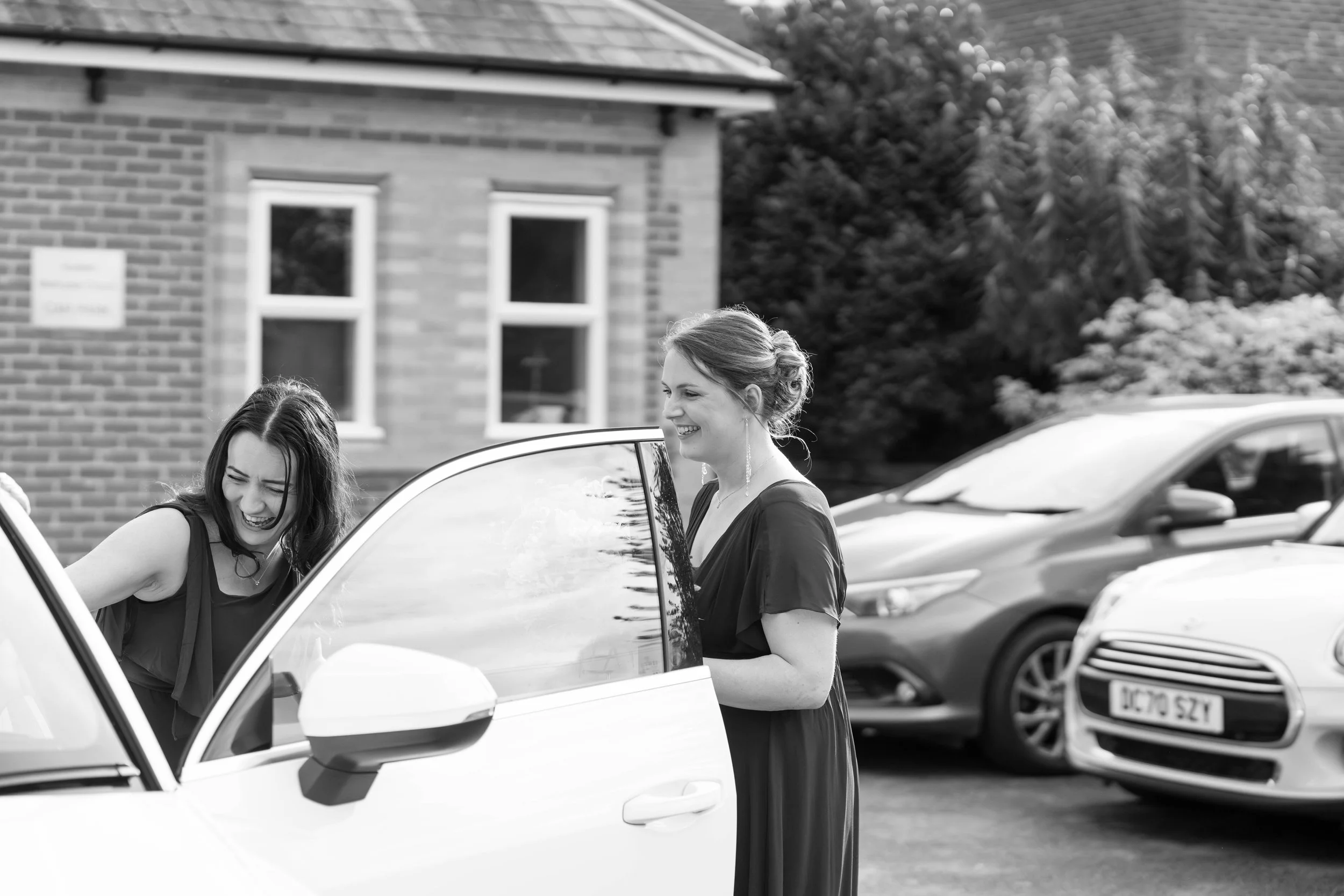 Two women laughing and smiling outside, one leaning into a car with the door open, in a parking lot with other cars and a brick building in the background.