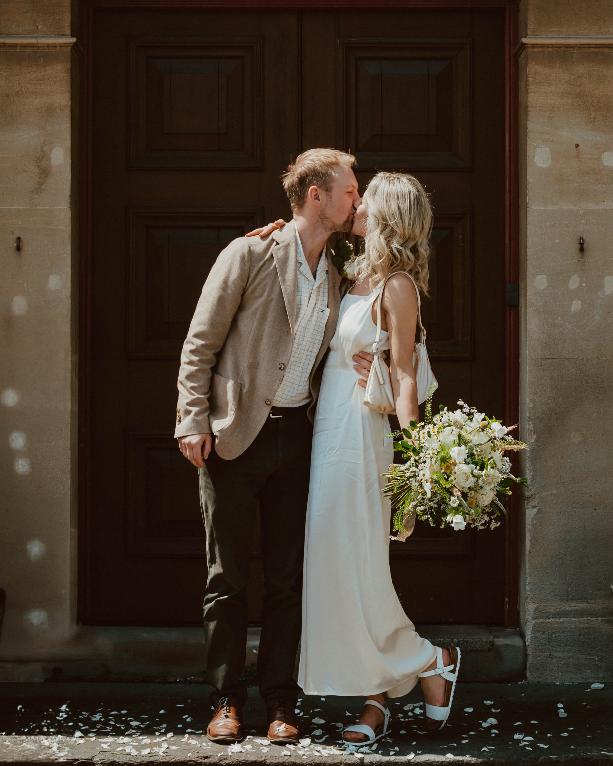 Couple kissing in front of a large wooden door, with a bouquet of white flowers, petals on the ground, and sunlight.