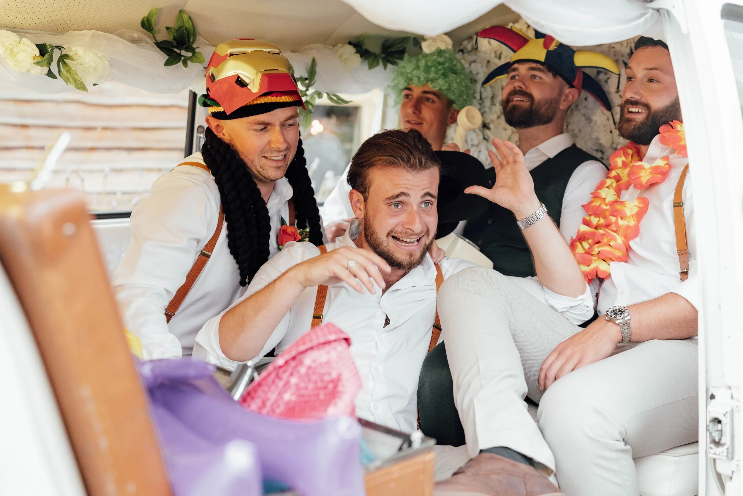A group of five men dressed casually with playful accessories, inside a decorated vehicle, celebrating and posing for a photo.