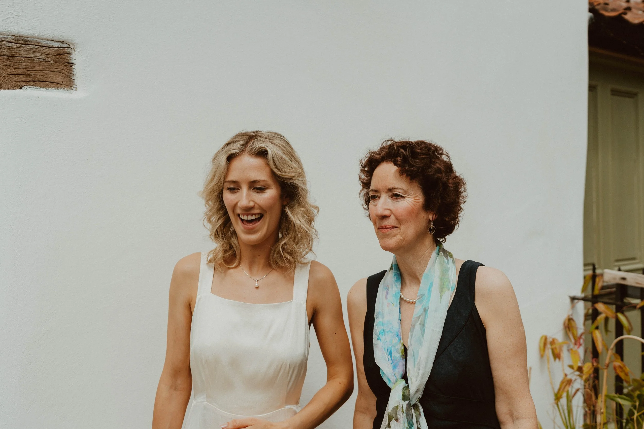 Two women standing and smiling against a white wall, one with blonde curly hair wearing a white dress, and the other with short curly hair wearing a black dress and a light-colored scarf.