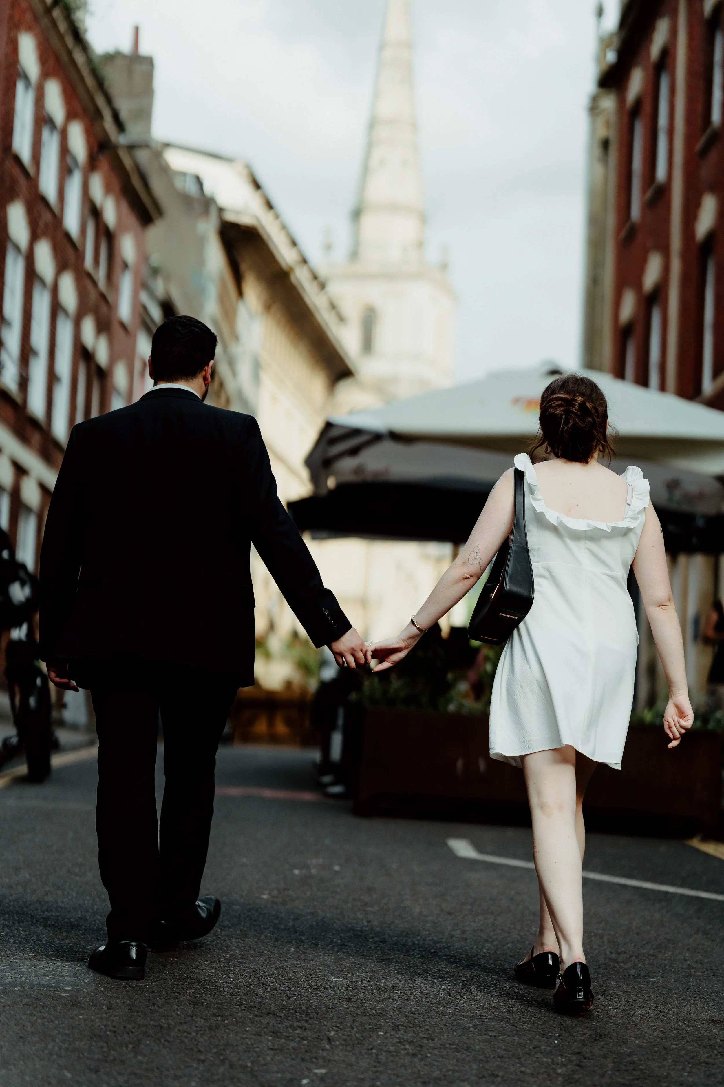 A man and woman holding hands while walking down a city street, seen from behind. The man is wearing a black suit, and the woman is in a white dress with a black bag over her shoulder.