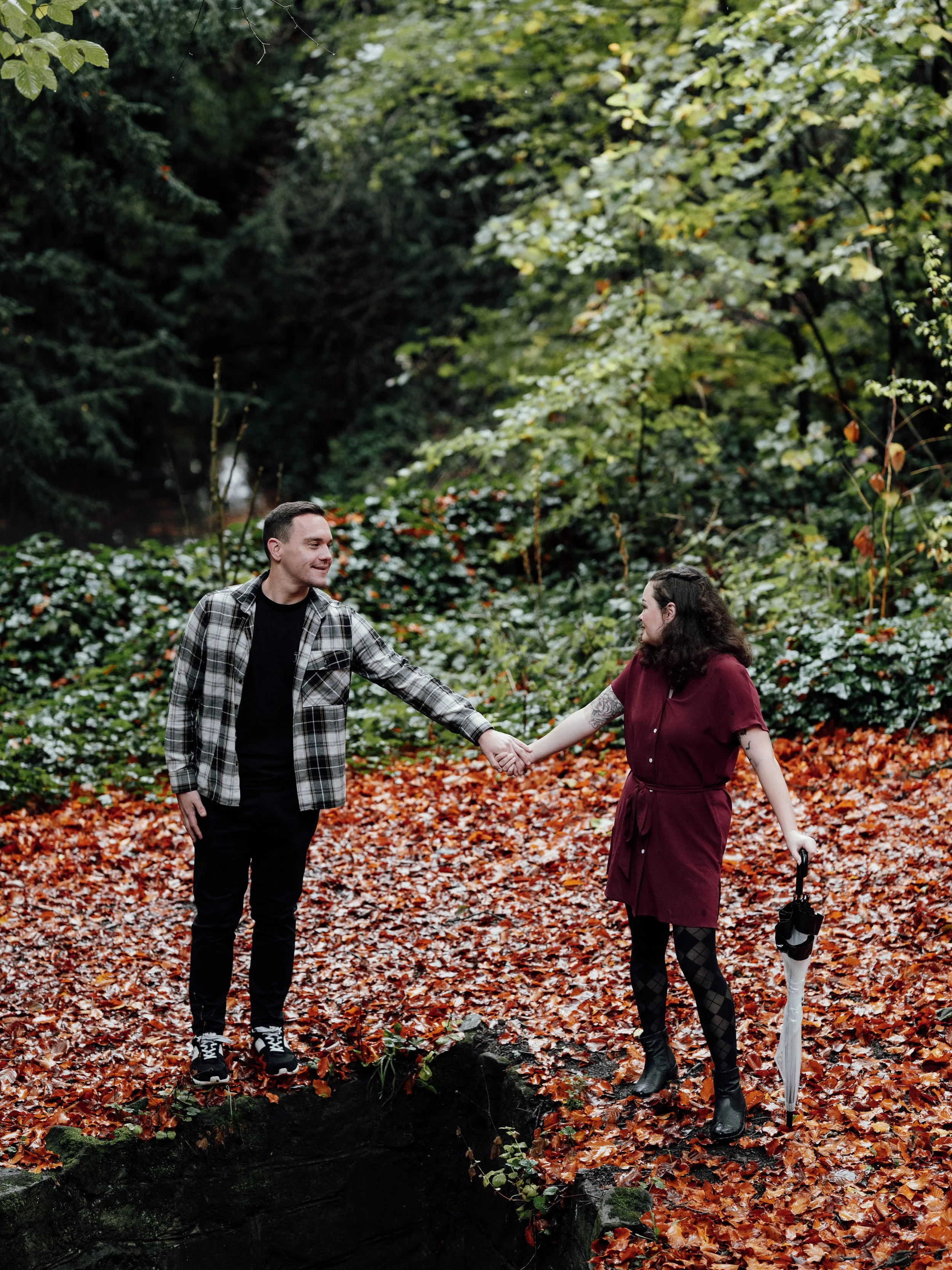A man and woman holding hands outdoors in a wooded area with fallen autumn leaves, trees, and greenery in the background, the woman holding an umbrella.