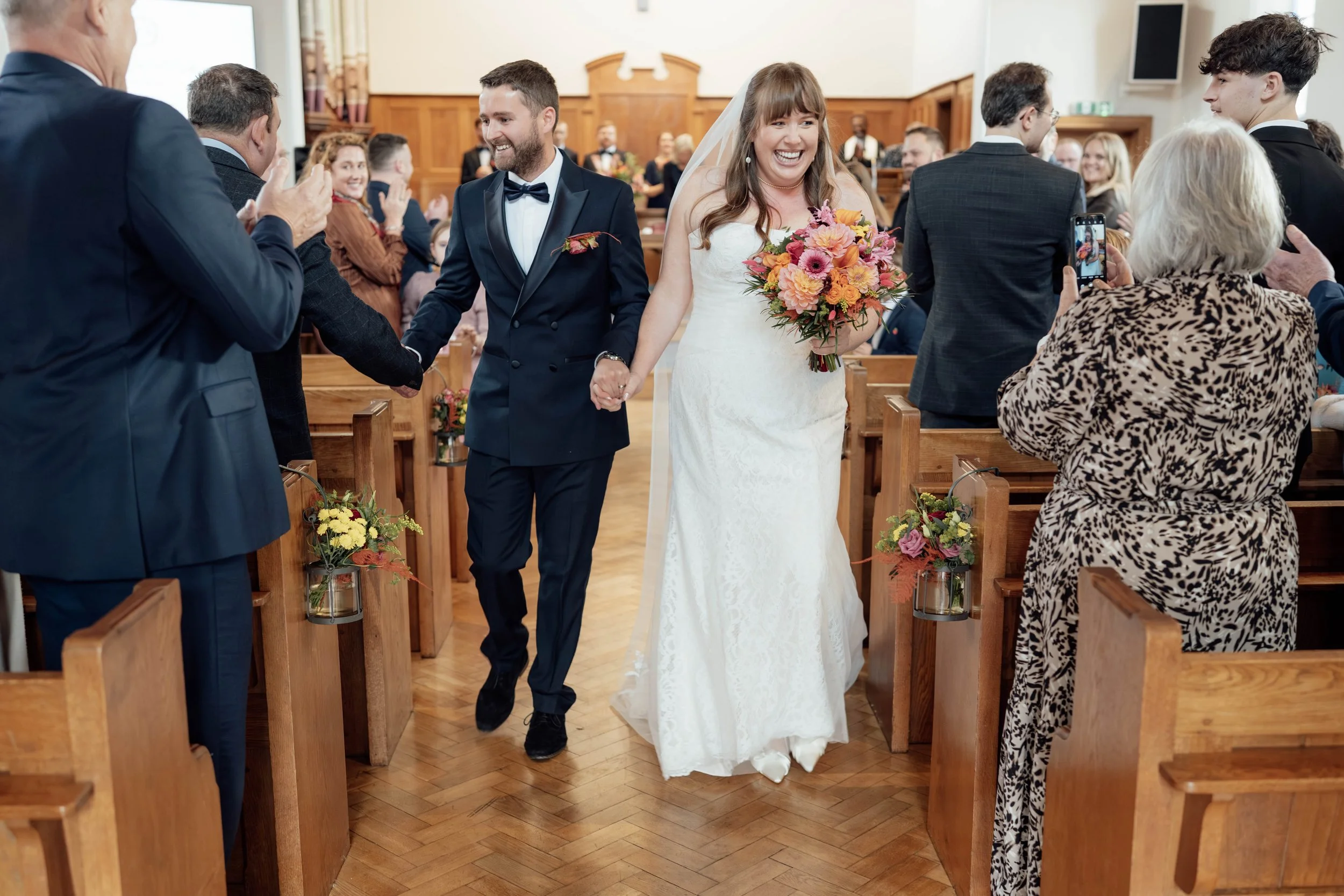 Bride and groom walking down the aisle holding hands, surrounded by guests taking photos at a wedding ceremony in a church.