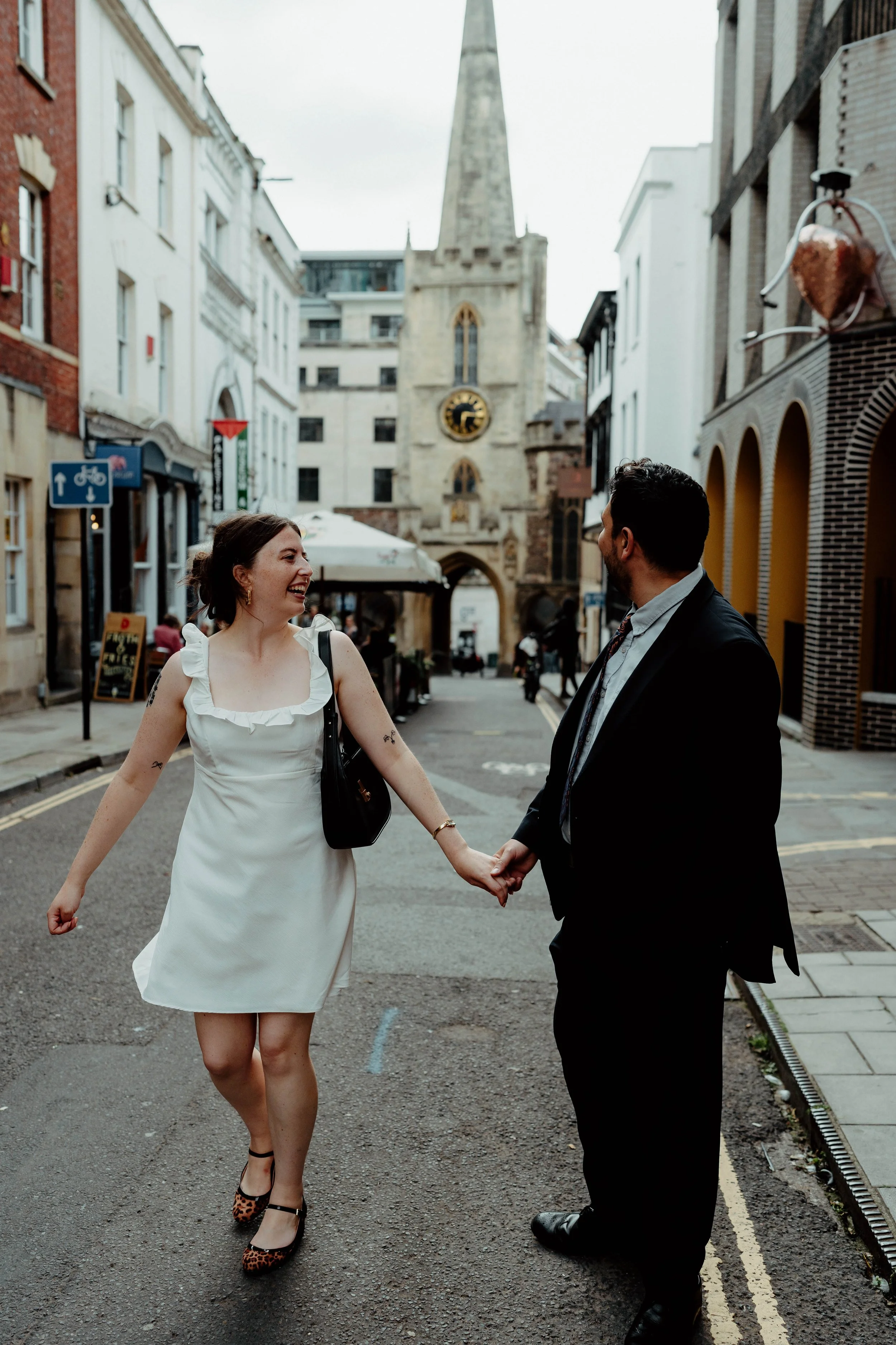 A woman and a man holding hands and smiling in a city street with historic buildings and a church tower with a clock in the background.