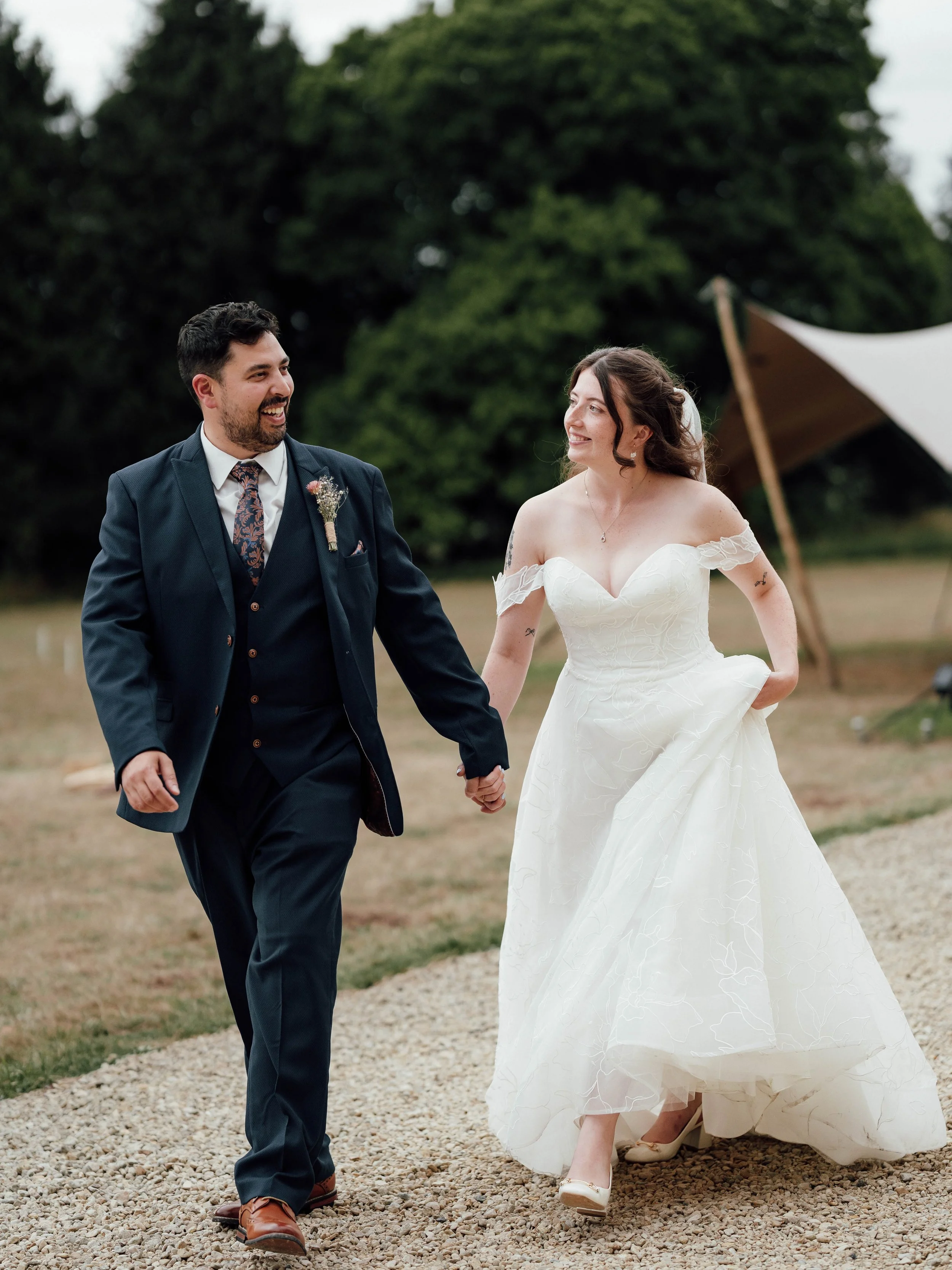A bride and groom happily walk hand in hand outdoors during their wedding, with trees and a tent in the background.