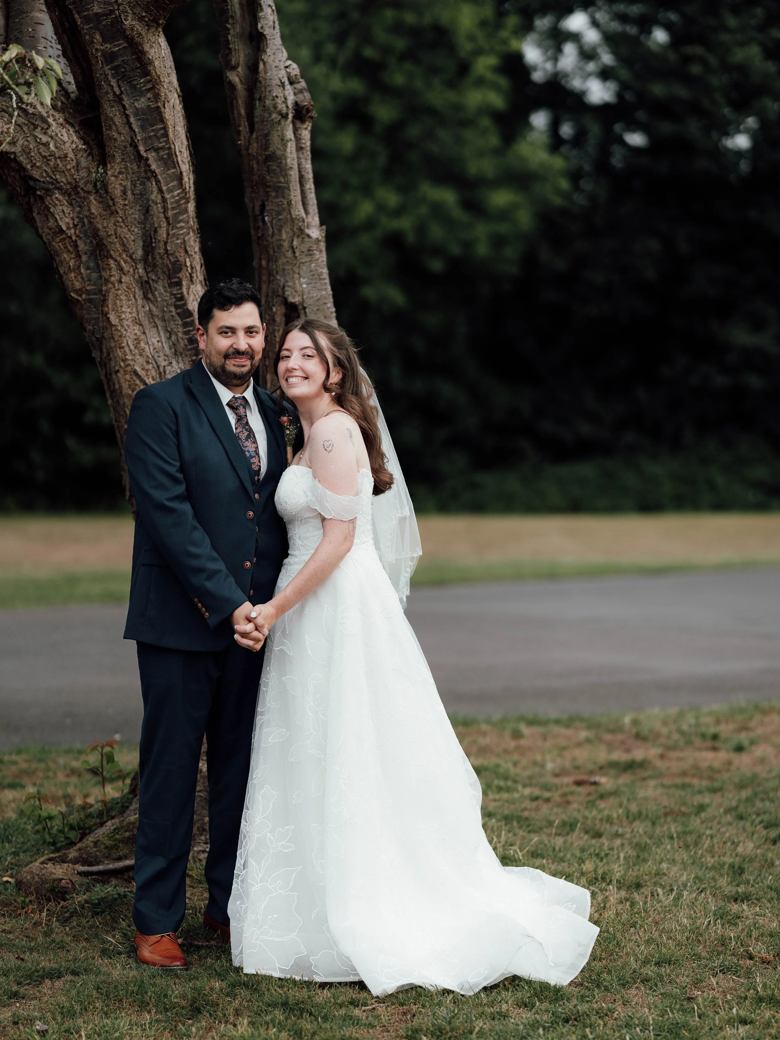 A bride and groom holding hands and smiling outdoors by a tree on their wedding day.