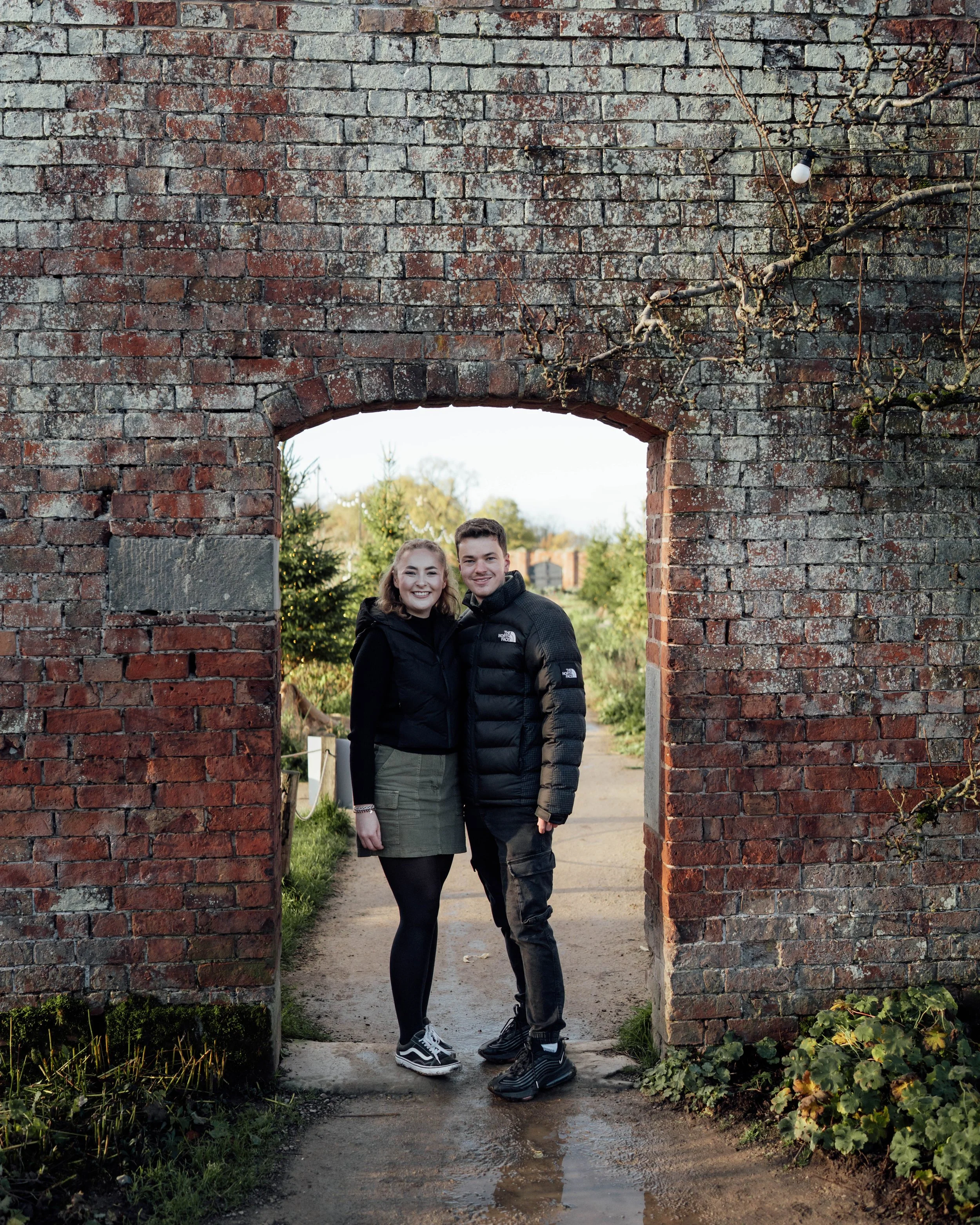 Smiling young couple standing in a brick archway outdoors, dressed warmly, with trees and a garden in the background.