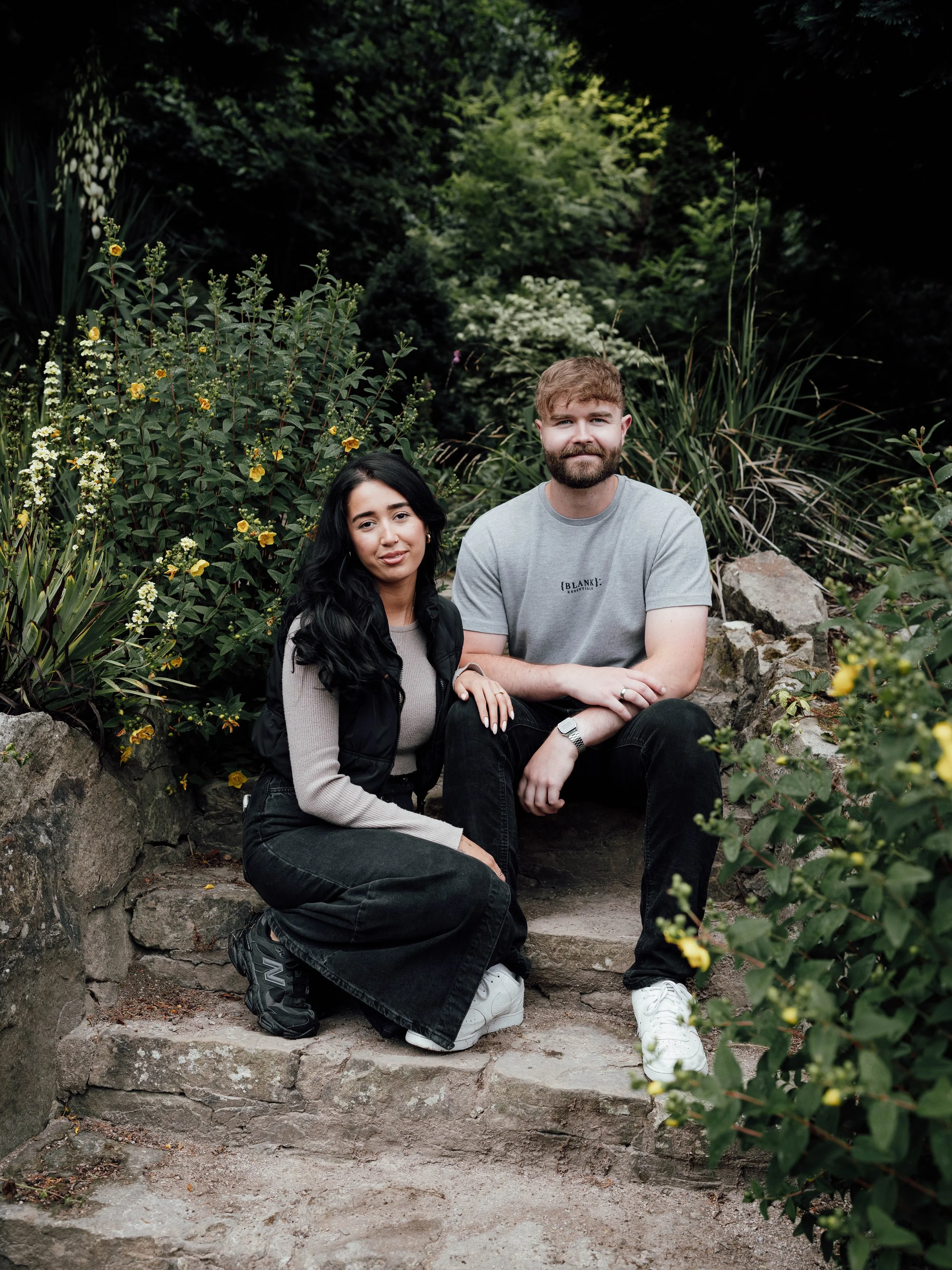 A young woman with long black hair and a young man with a beard and short hair sitting on stone steps in a garden with lush greenery and yellow flowers.