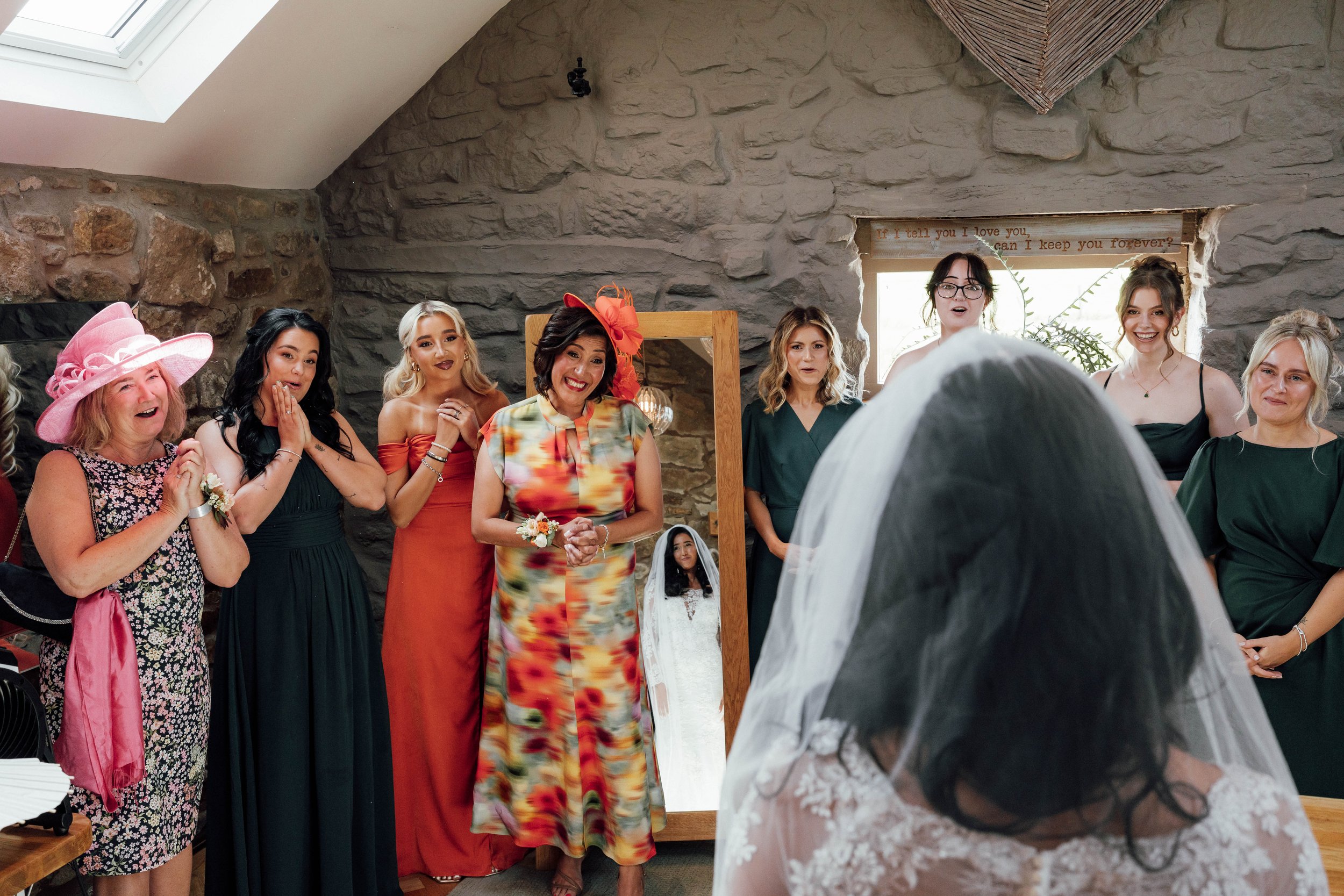 A group of women dressed in colorful and elegant attire are gathered indoors in surprise and joy, facing a woman in a white wedding dress with a veil. The scene appears to be at a wedding-related event, possibly a bridal shower or wedding preparation