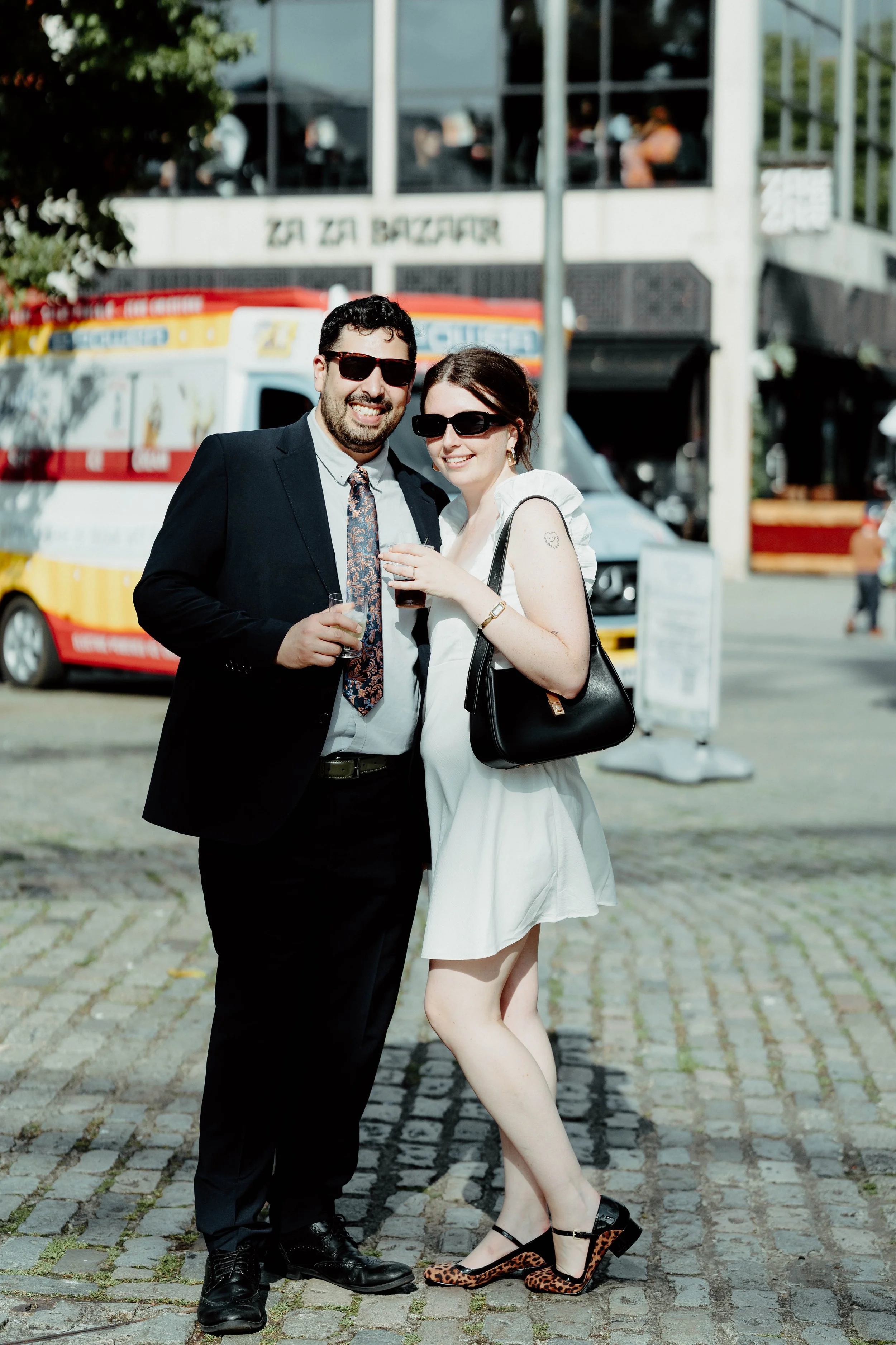 A man and woman standing outdoors on a cobblestone street, smiling and holding drinks, with an ice cream truck and a building with large windows behind them.