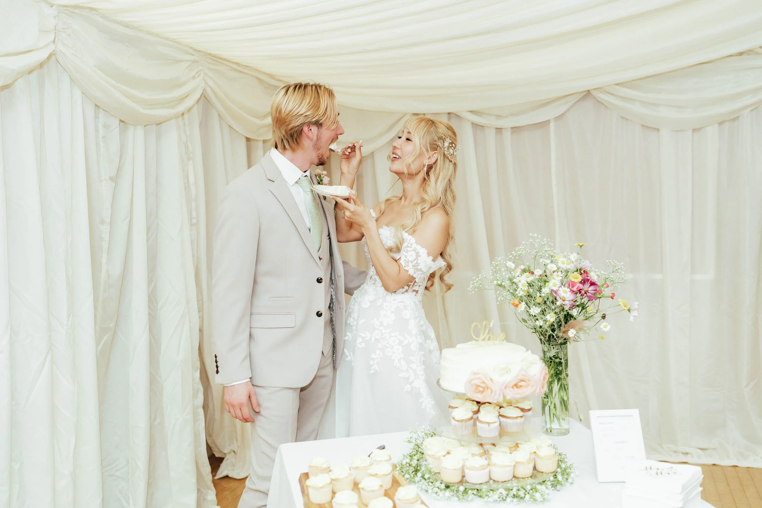 Bride and groom at wedding reception, feeding each other cake in a decorated venue with a table of cupcakes, flowers, and a cake.