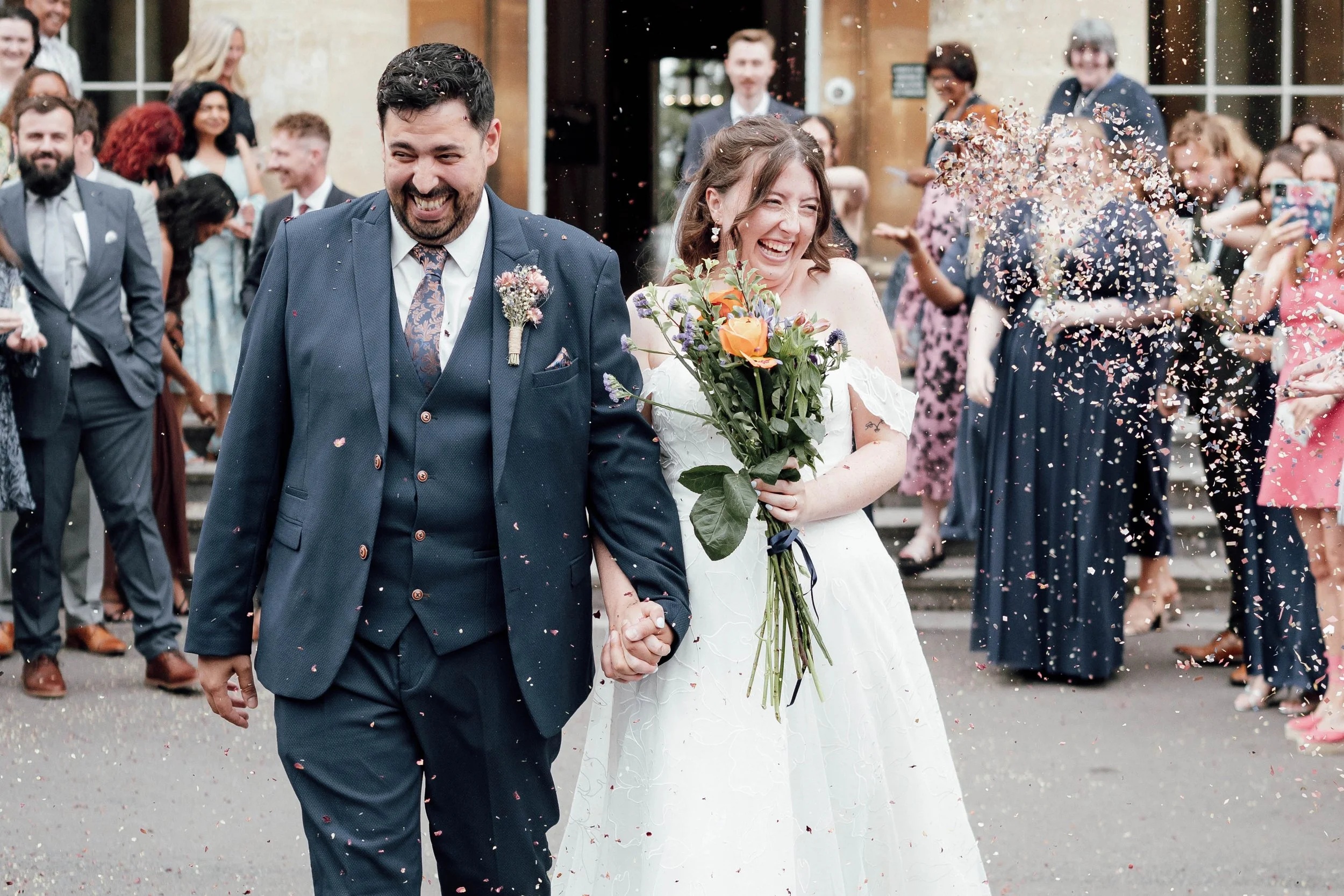 A newly married couple holding hands and smiling as confetti falls around them, with wedding guests cheering in the background.