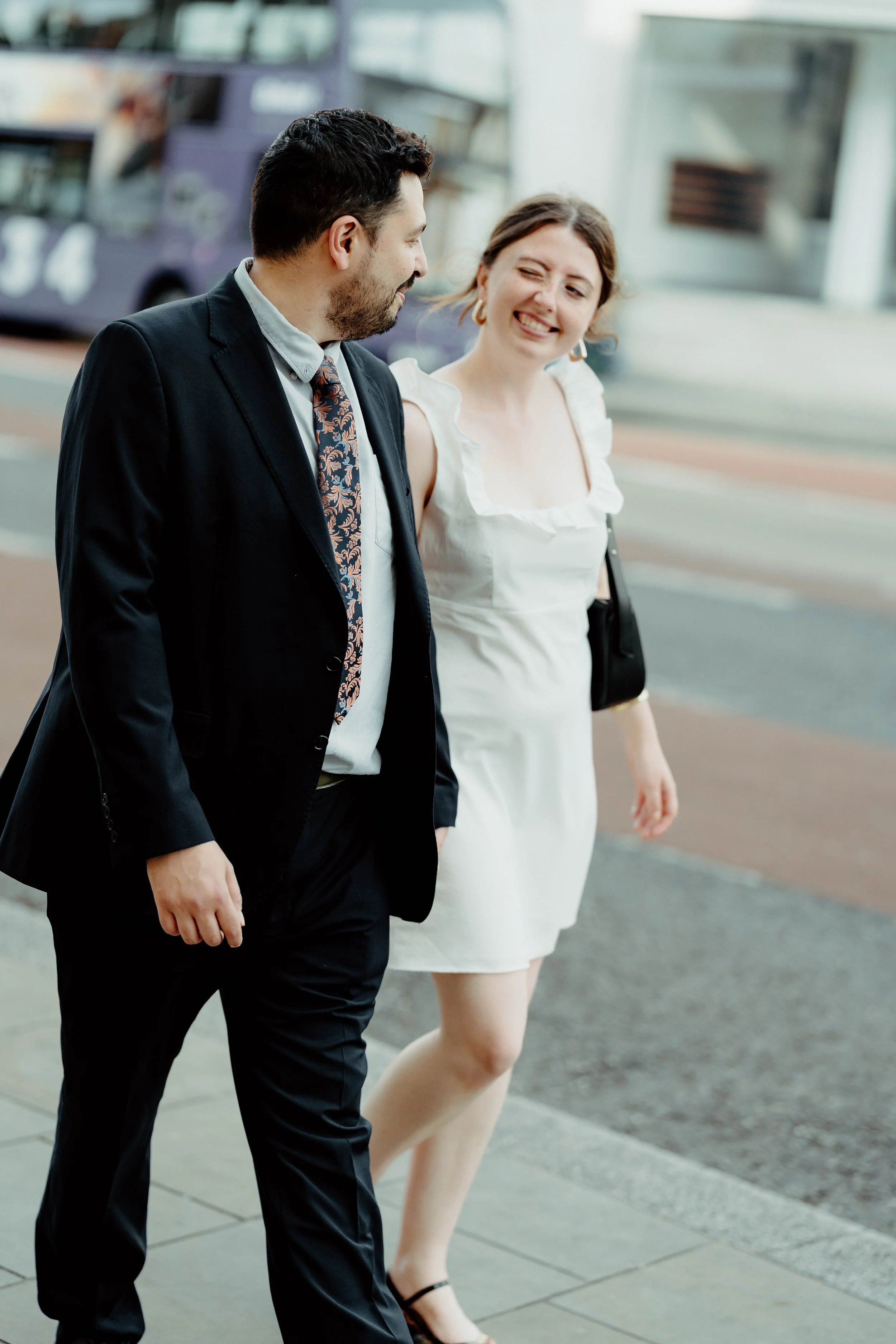 A man and woman walking together on a city sidewalk, smiling and engaged in conversation, with a city bus in the background.