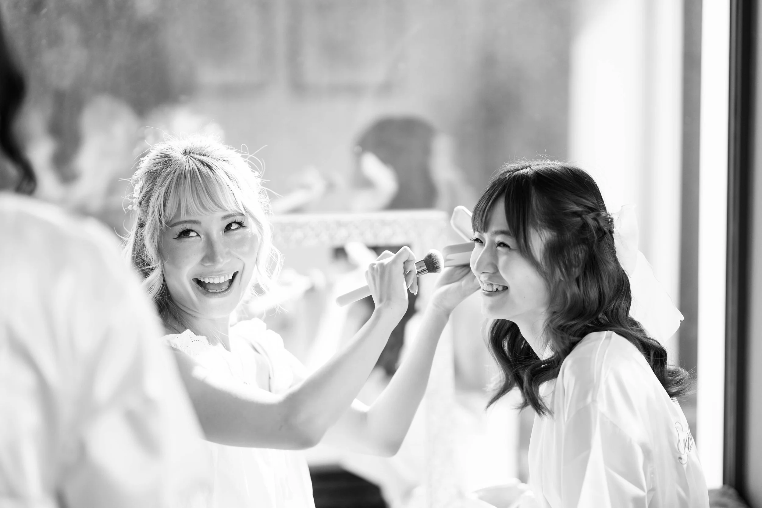 Women in a makeup room, one person applying makeup to another, both smiling.