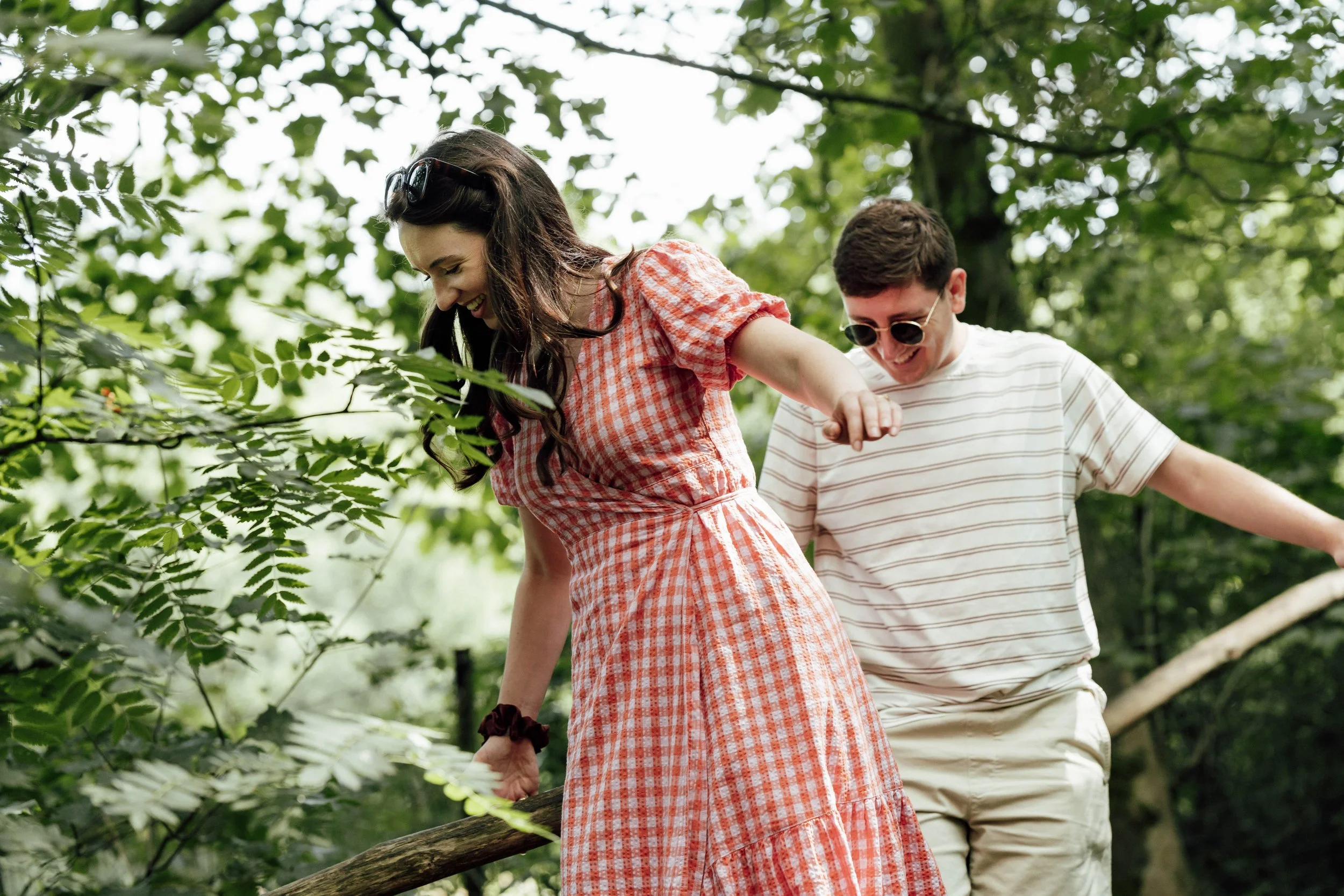 A young woman and man are smiling and walking through a wooded park, holding onto a wooden railing, enjoying a sunny day.