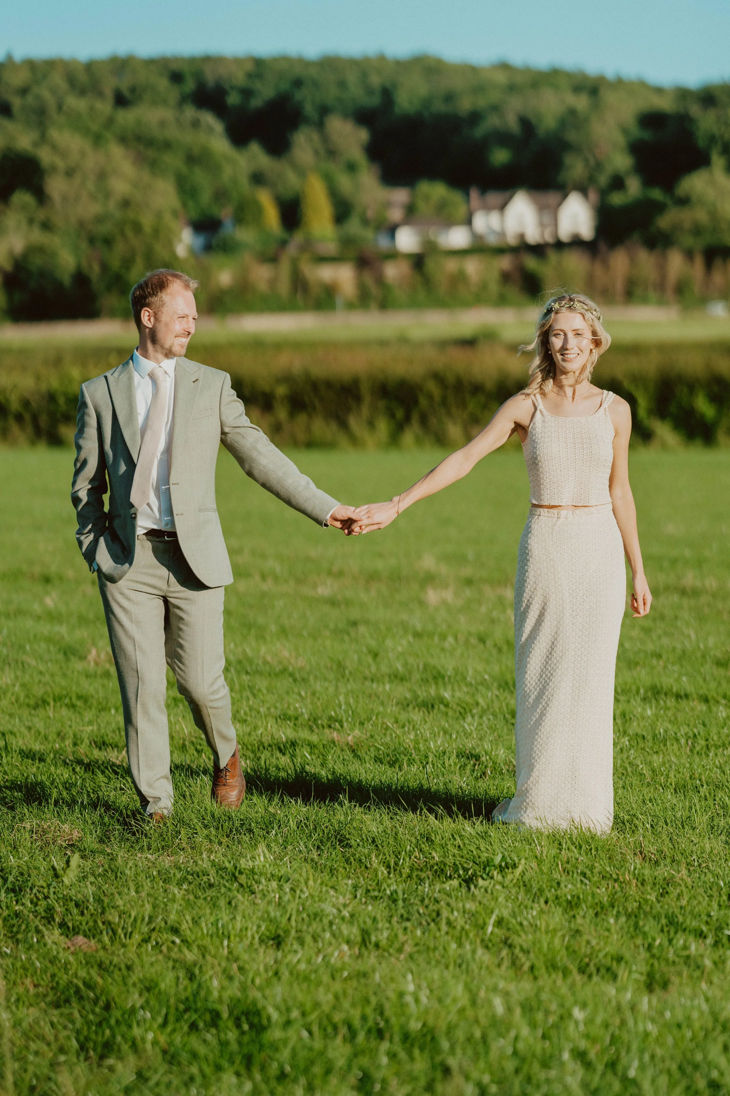 A couple holding hands outdoors on a grassy field, with trees and houses in the background on a sunny day.