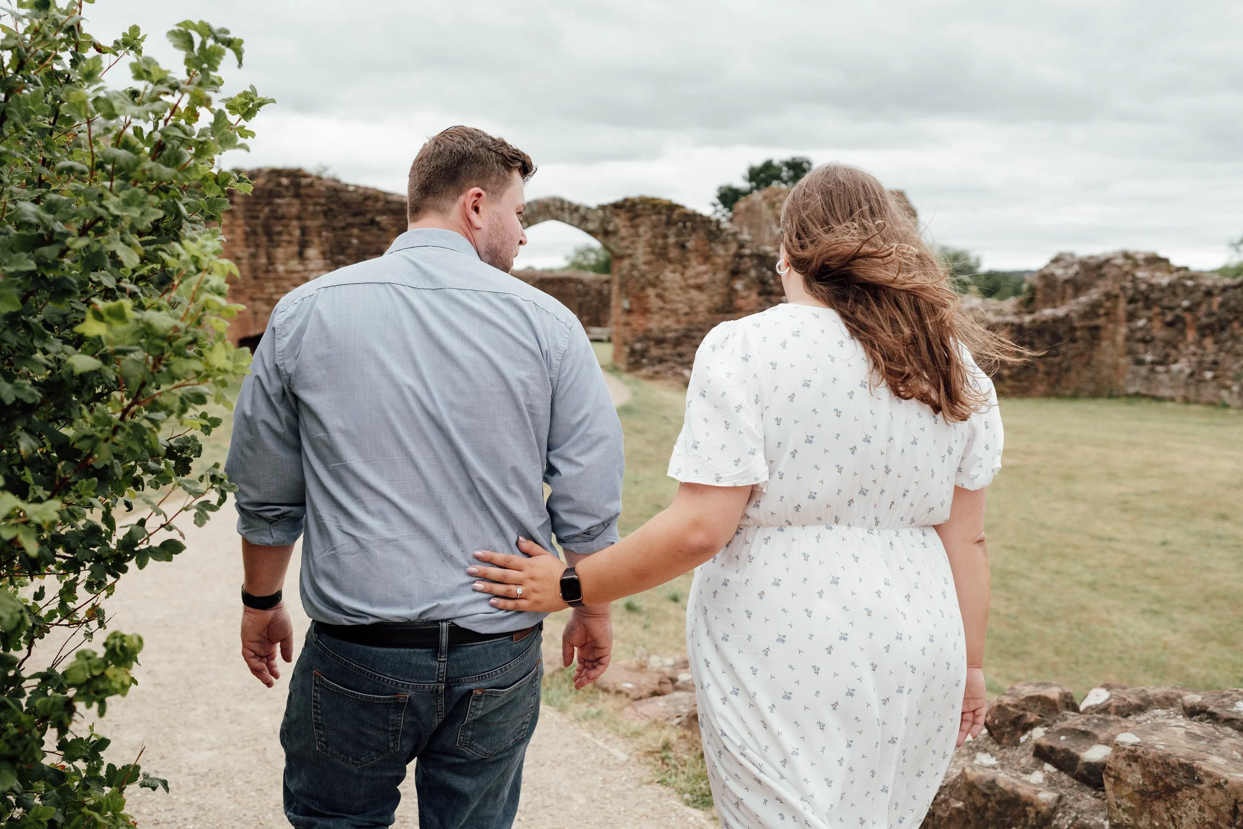 A couple walking together outdoors, holding hands, with ancient brick ruins in the background, overcast sky.