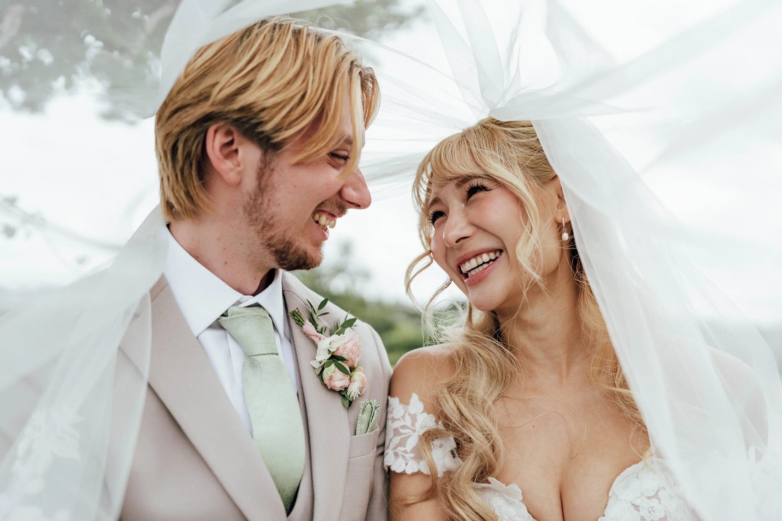 A smiling bride and groom under a veil, sharing a joyful moment on their wedding day outdoors.