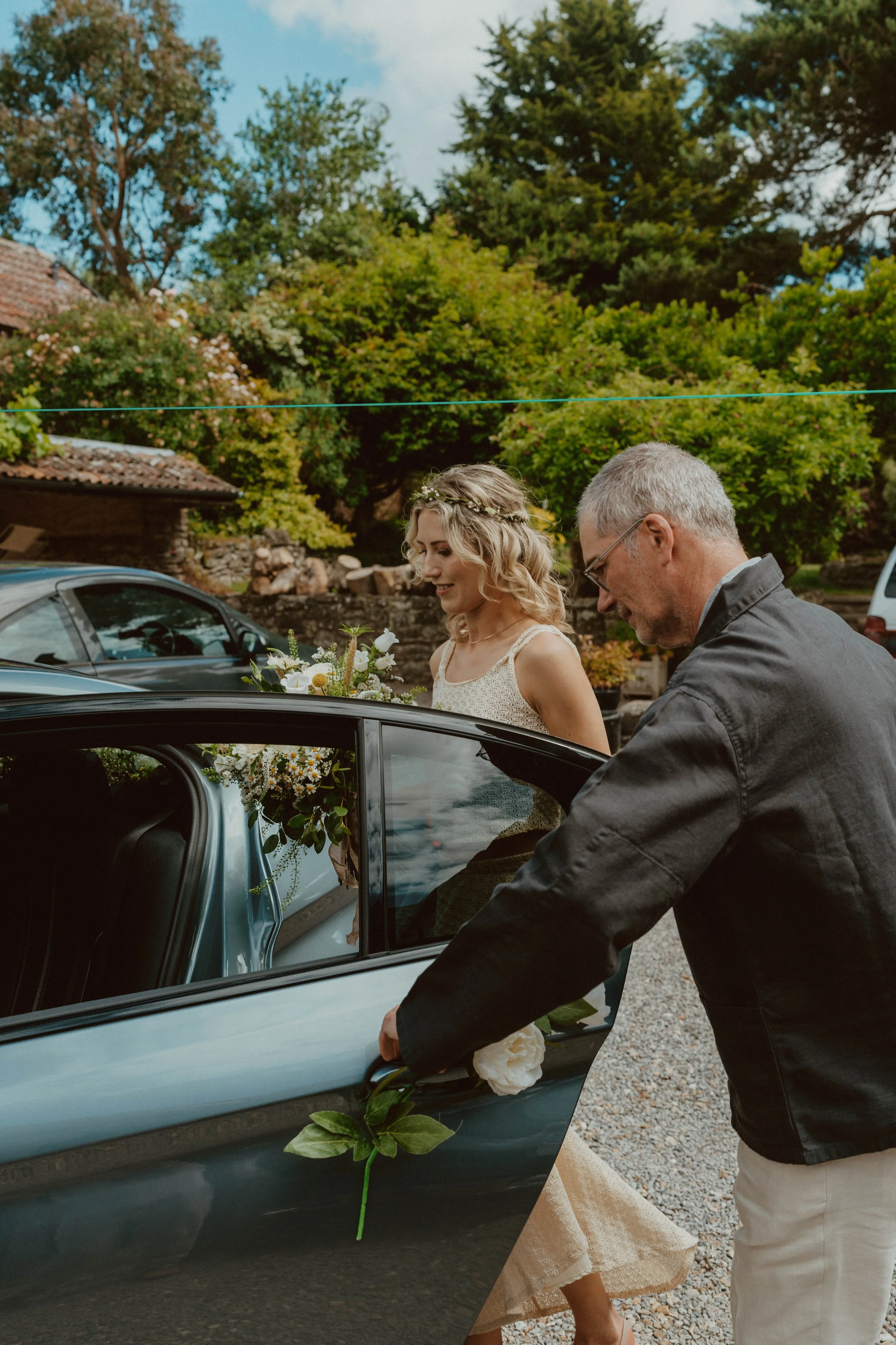 A woman in a white dress and a floral crown, holding a bouquet, getting into a car at an outdoor wedding. A man is opening the car door with a white flower in his hand.