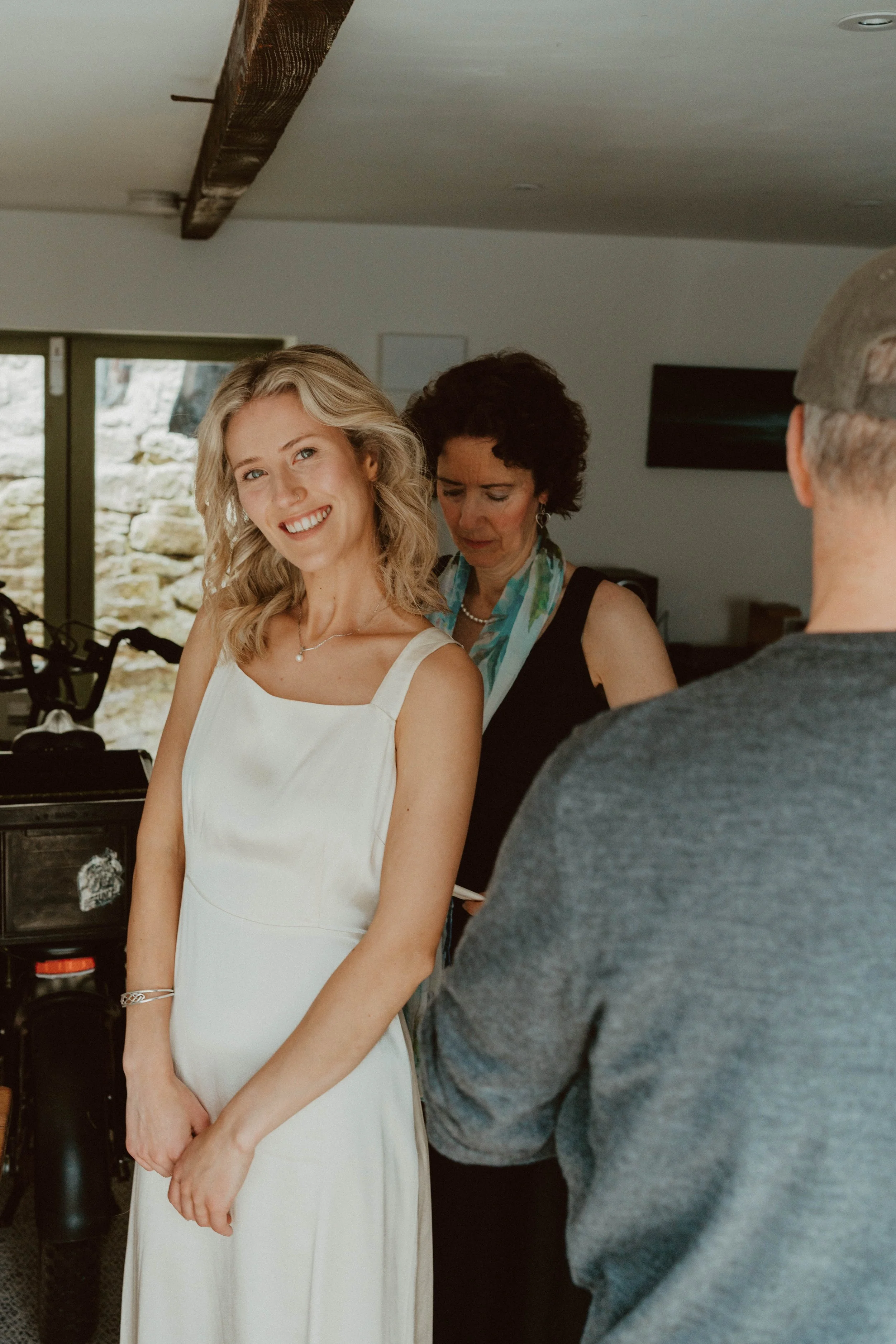 A young woman in a white dress smiling at the camera while standing indoors, with an older woman behind her looking down, and a man in gray clothing with his back to the camera in the foreground.
