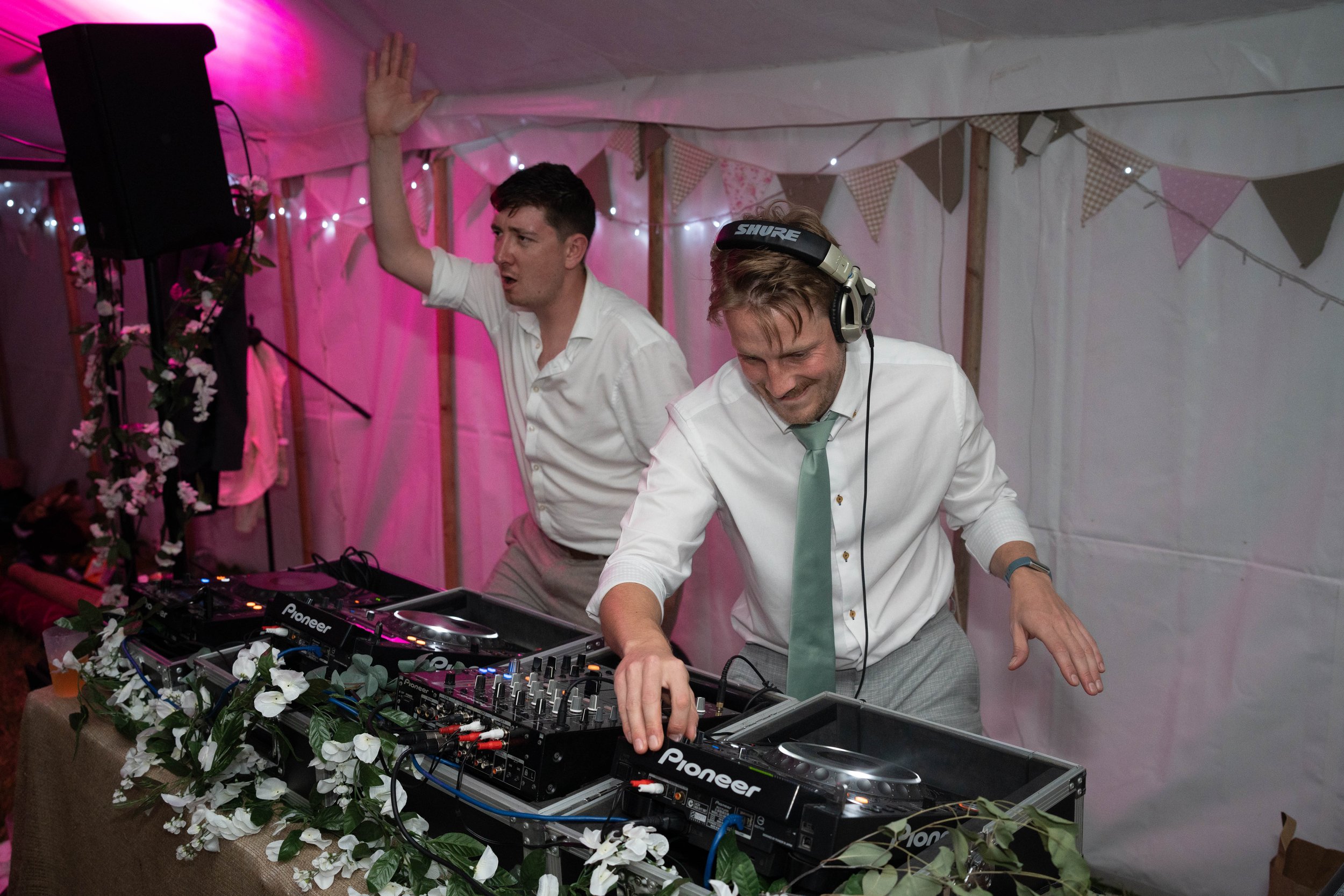 Two men, one DJ with headphones and a light blue tie, and another man raising his hand, are partying with DJ equipment decorated with white flowers inside a pink-lit tent.