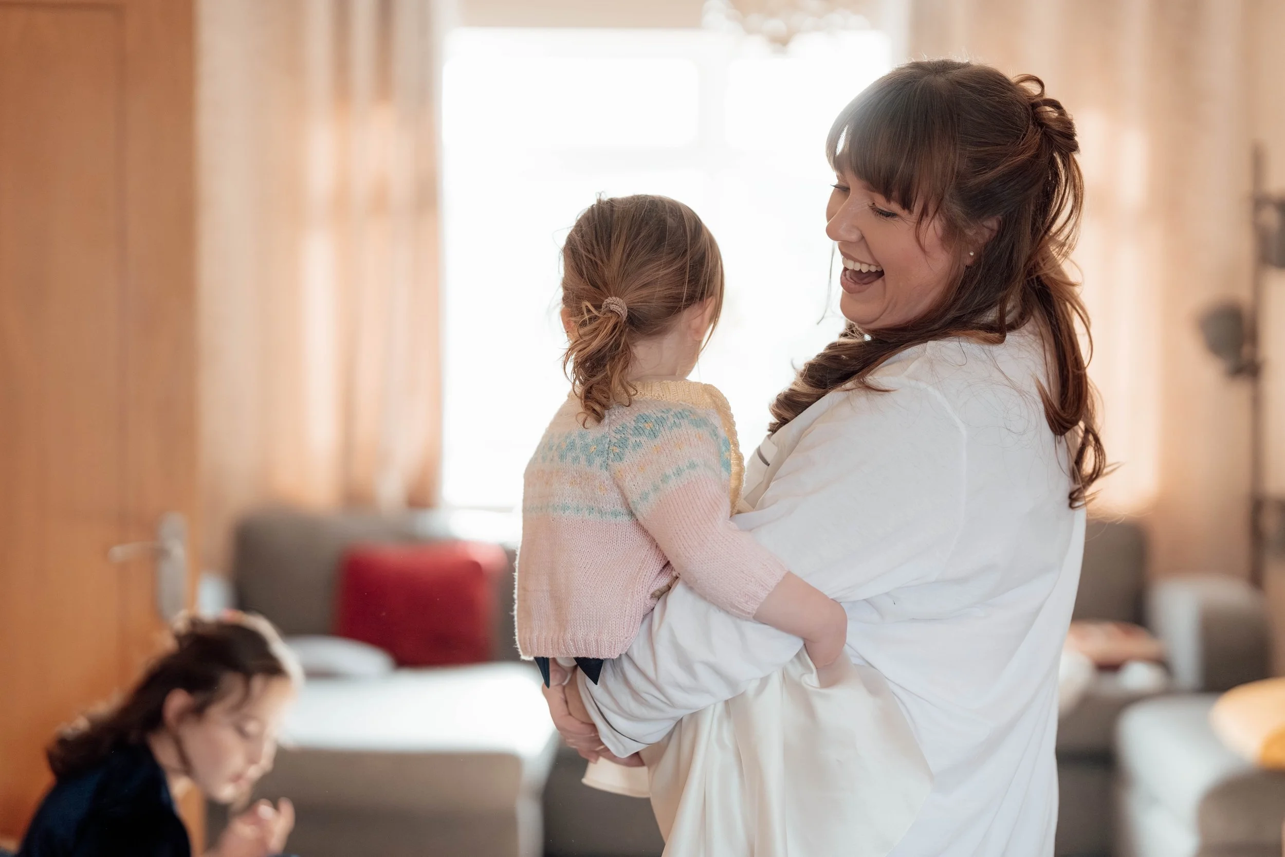A woman holding a young girl, both smiling and looking at each other in a cozy living room with sunlight coming through a window.