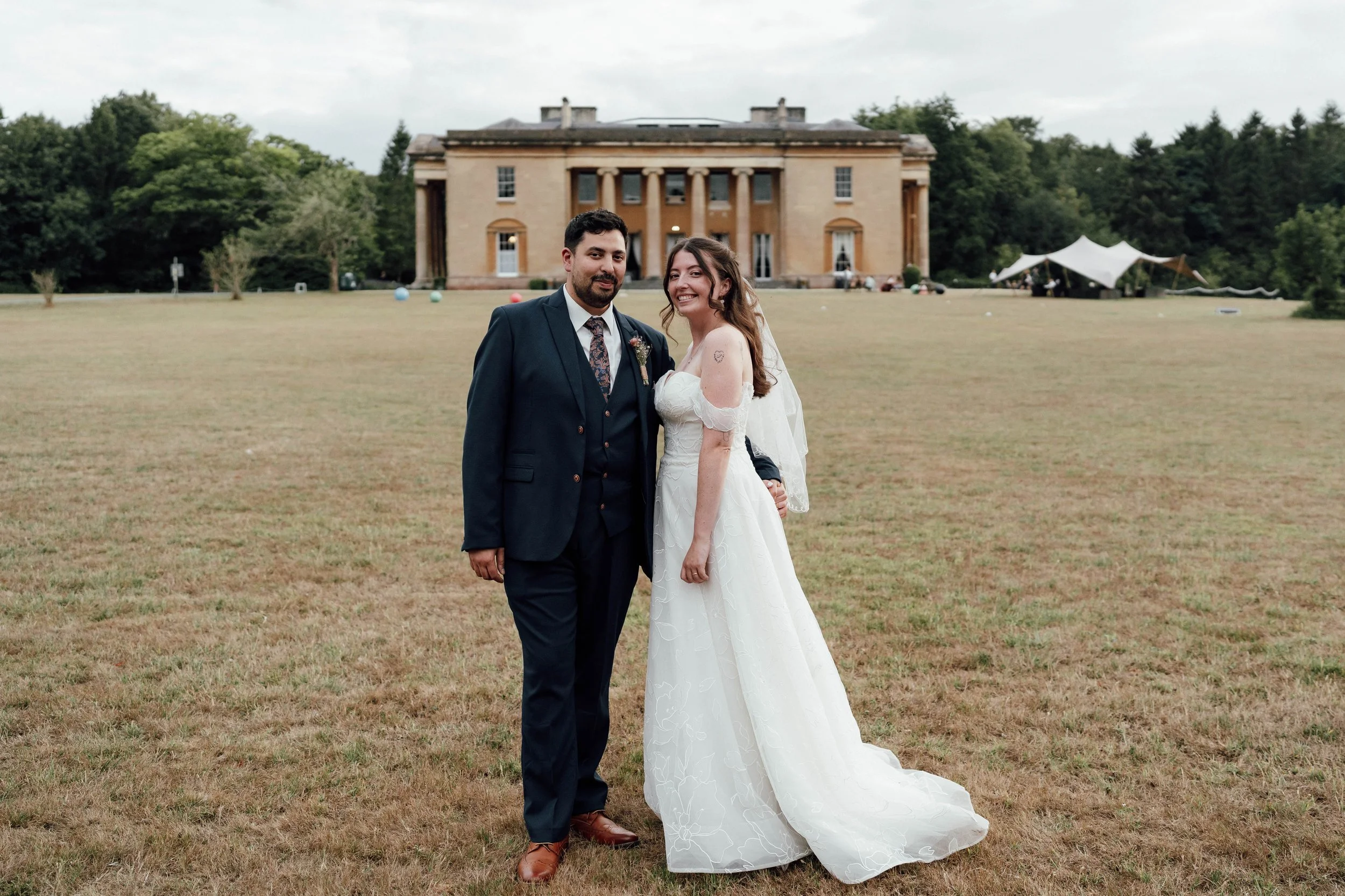 A bride in a white wedding dress and a groom in a dark suit standing on a grassy field in front of a large beige house with columns. The bride is smiling and has her arm around the groom, who is also smiling. There is a large white tent in the backgr