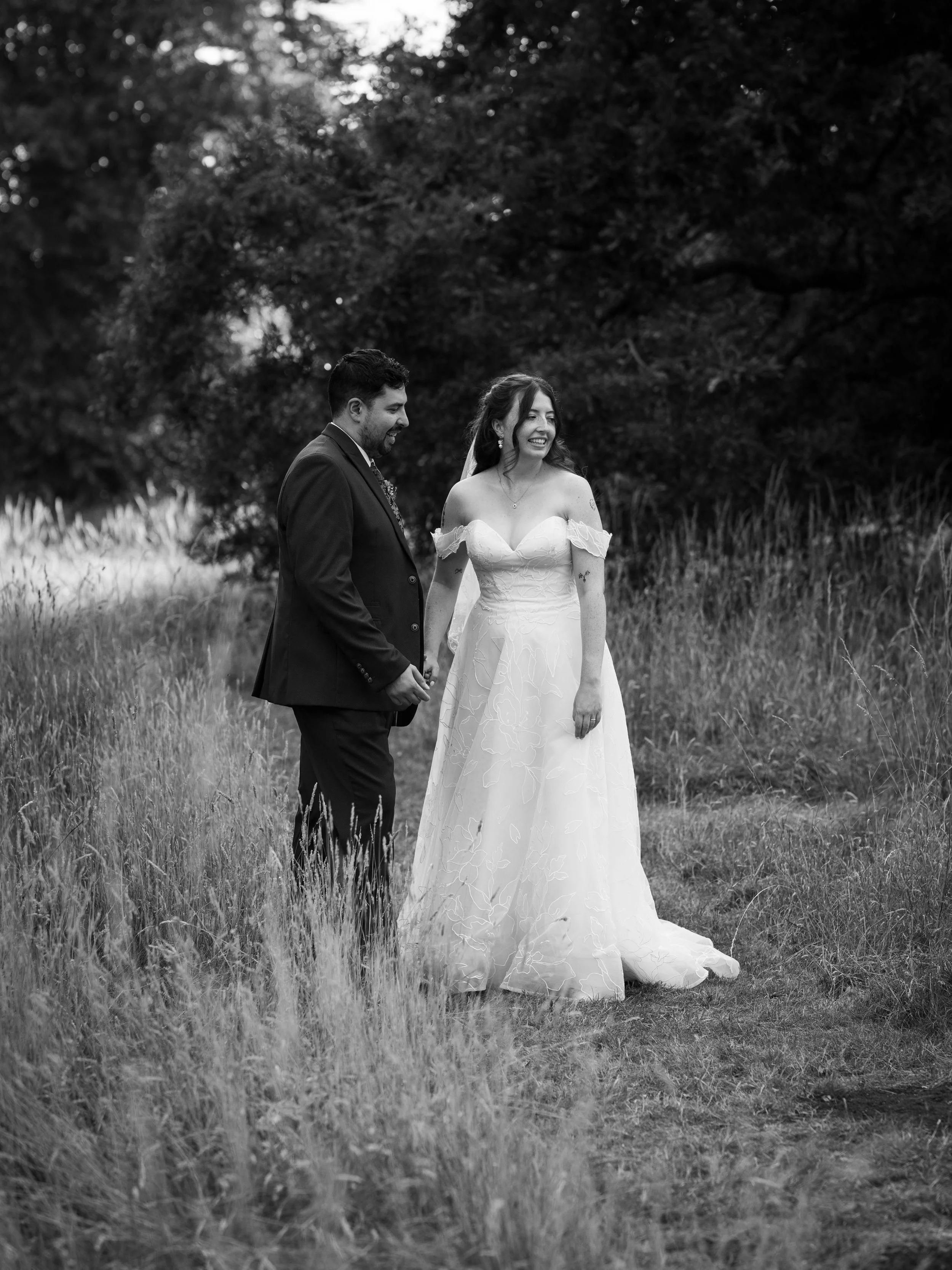 Black and white photo of a bride and groom standing outdoors on a grassy field, holding hands and smiling, with trees in the background.