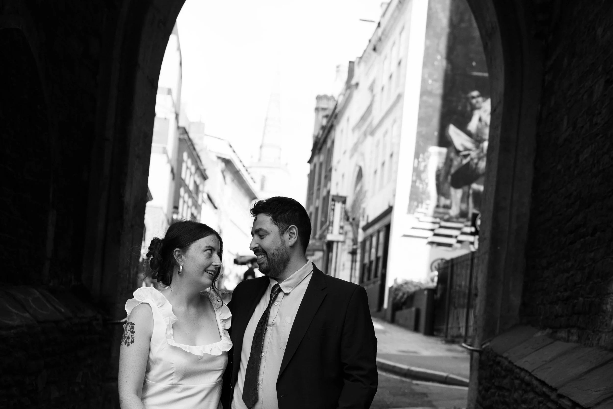 A man and woman smiling and looking at each other in an urban setting, walking under an archway. The woman has a tattoo on her upper arm and wears a ruffled sleeveless top. The man is dressed in a suit with a tie.