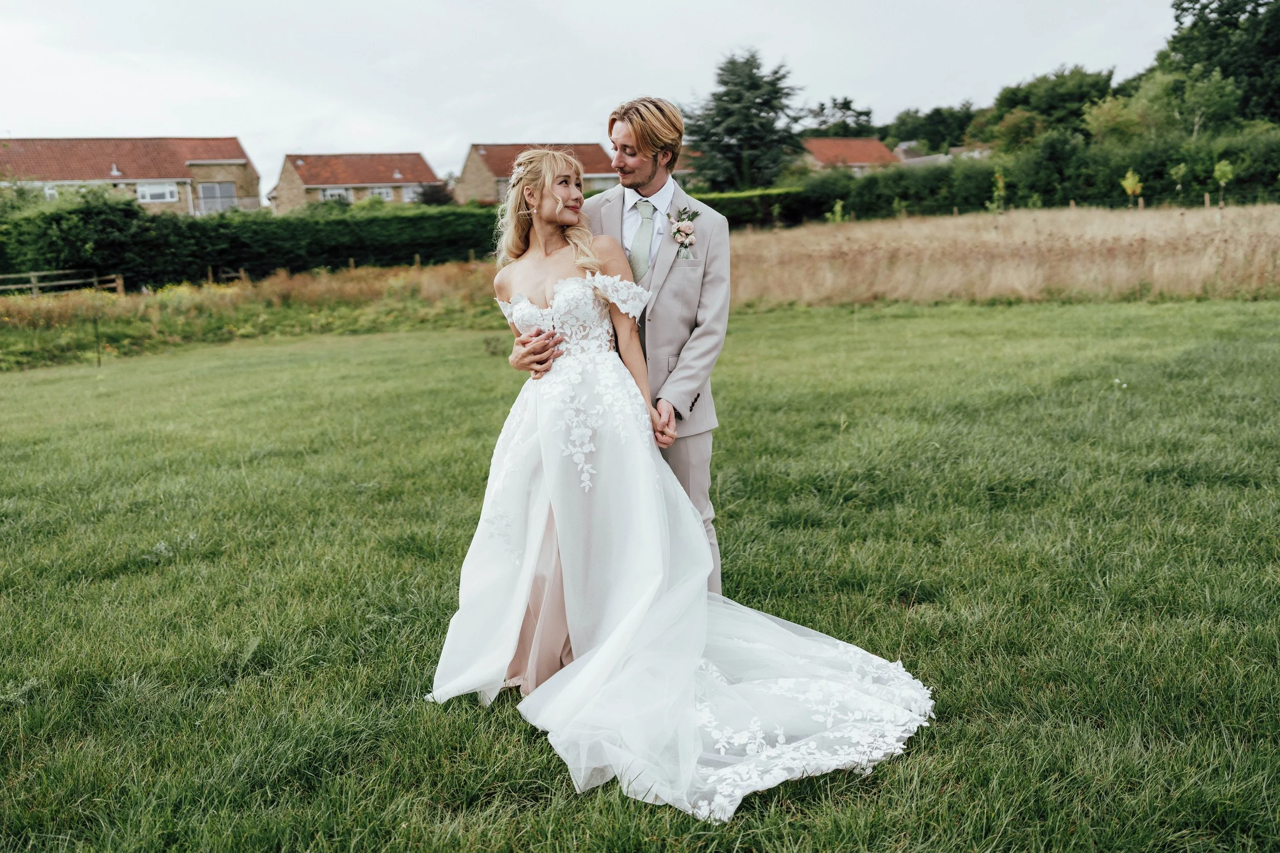 Bride and groom standing on green field, holding hands, with houses and trees in the background, during daytime.