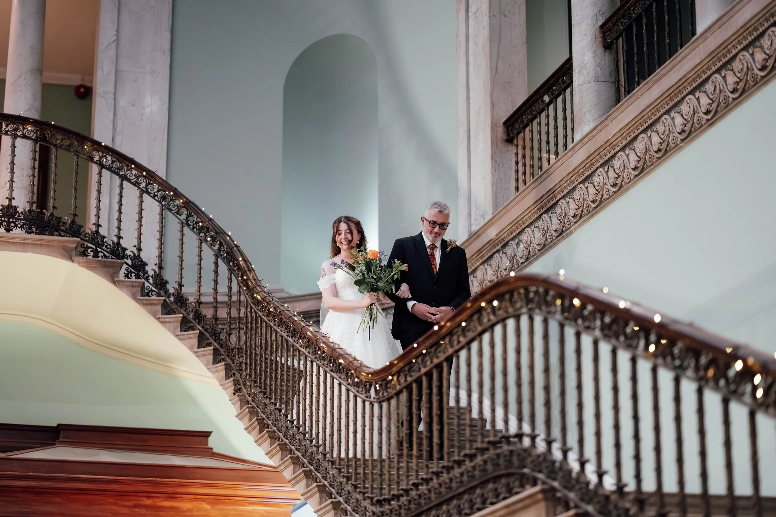 A bride in a white wedding dress holding a bouquet of flowers walking down a grand staircase with a man in a black suit, likely her father, during a wedding or special event in an elegant building with ornate railing and marble columns.