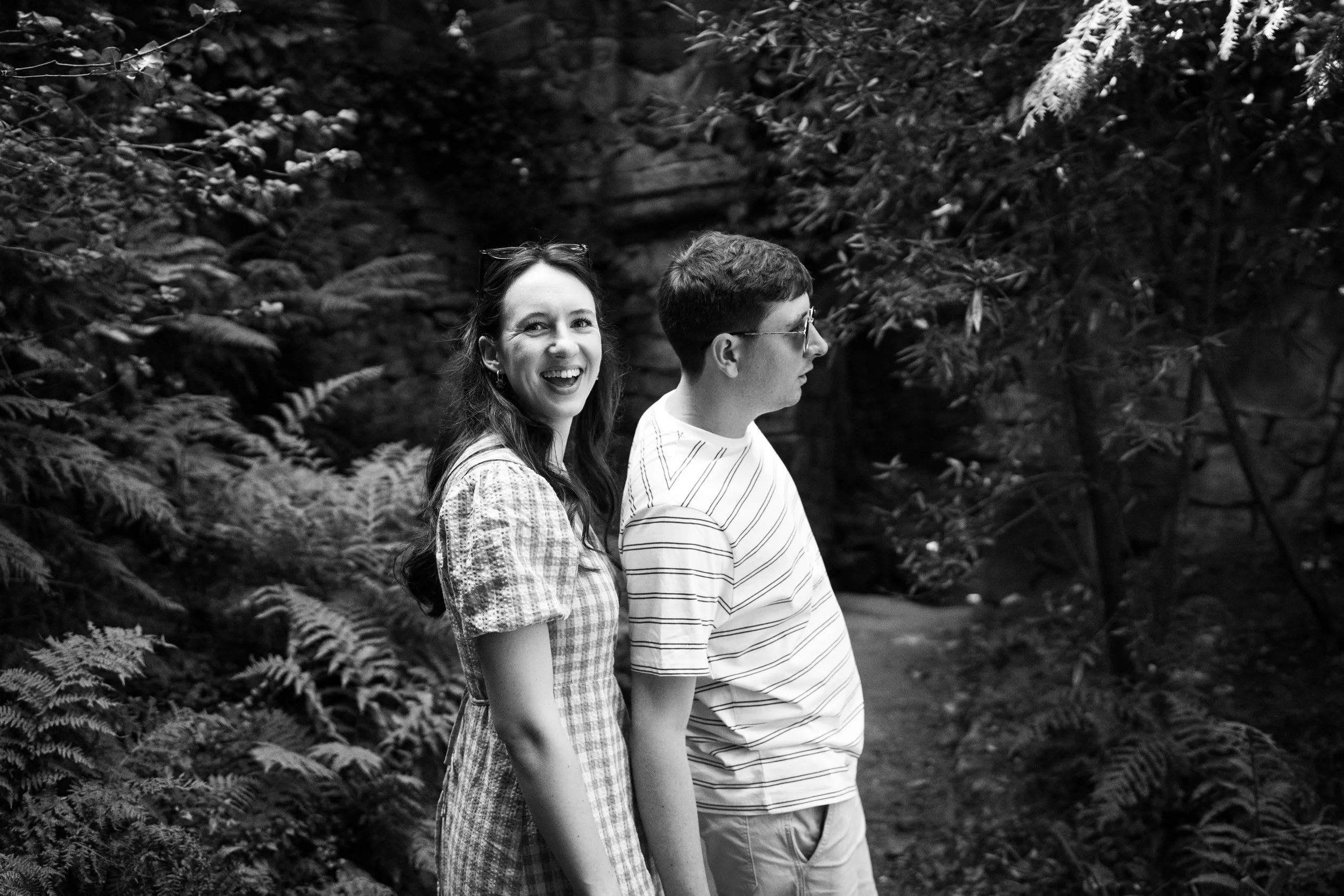 A smiling woman and a man in sunglasses standing outdoors in a wooded area.