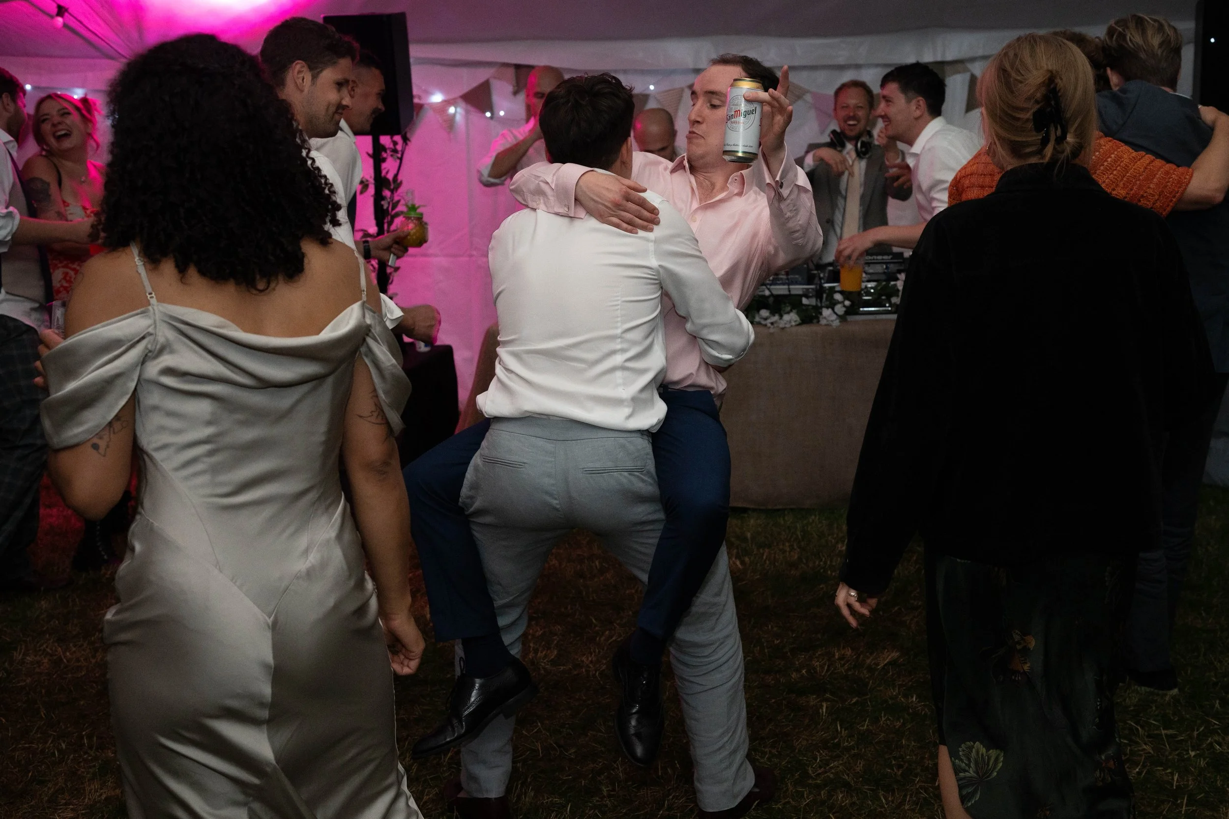 People dancing and celebrating at a party or wedding reception, with some hugging and holding beverages, in a decorated tent with string lights.