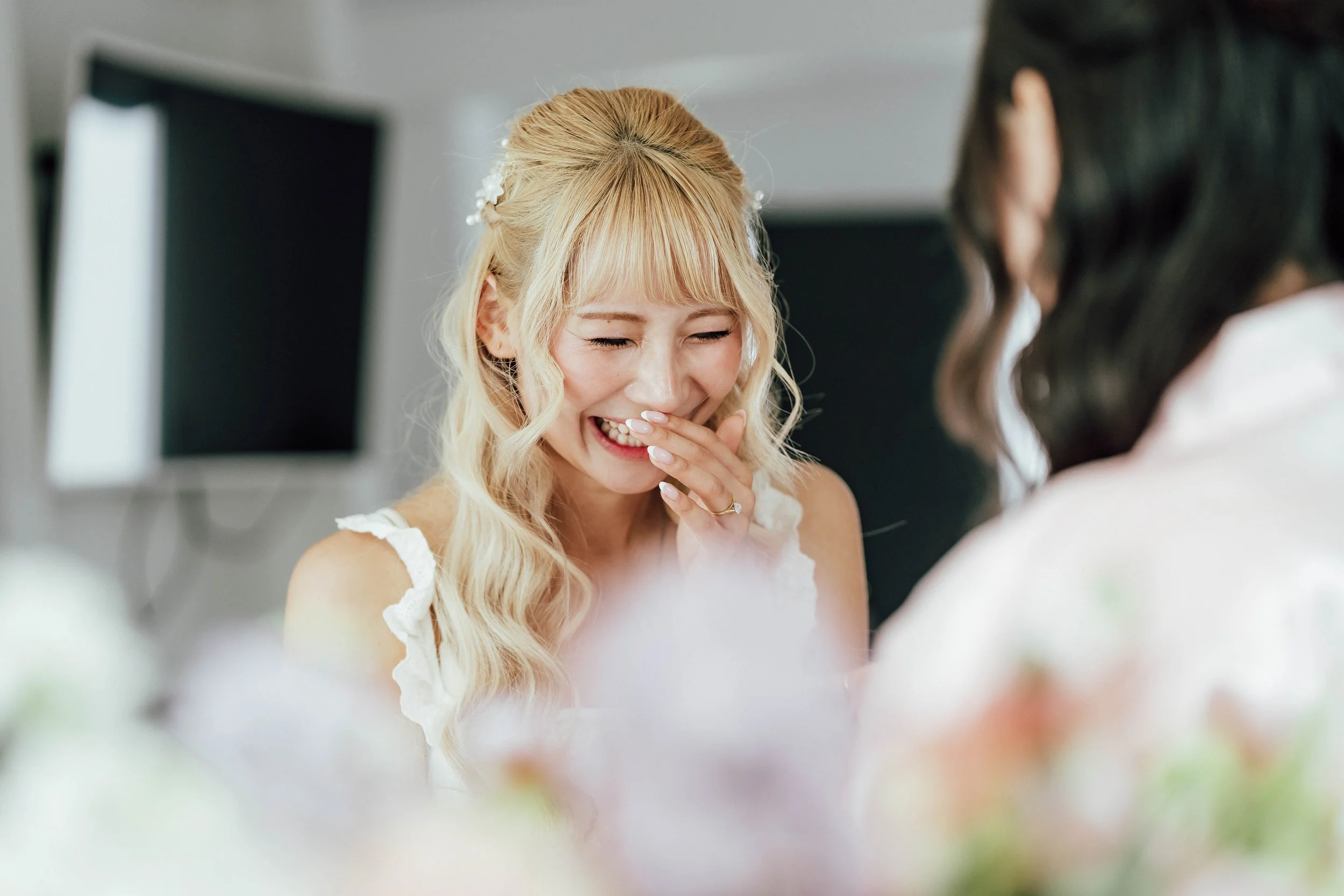 A blonde woman laughing and covering her mouth with her hand, wearing a white dress, in a joyful moment indoors.