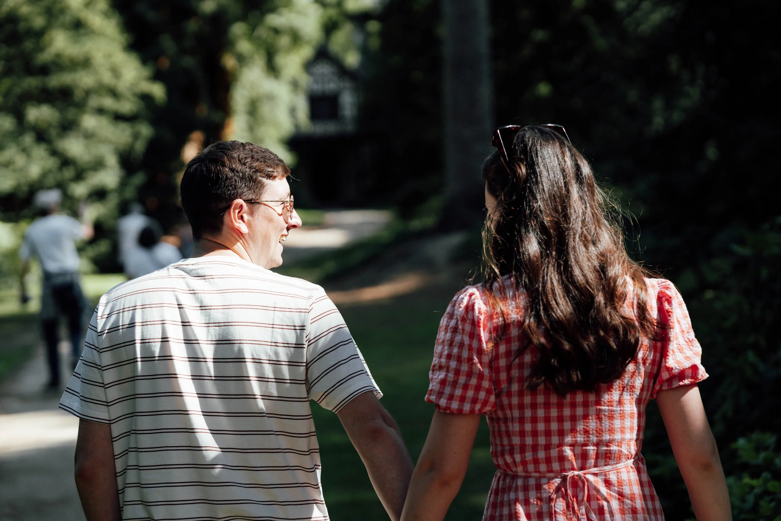 A man and woman walking hand in hand in a park on a sunny day, seen from behind.