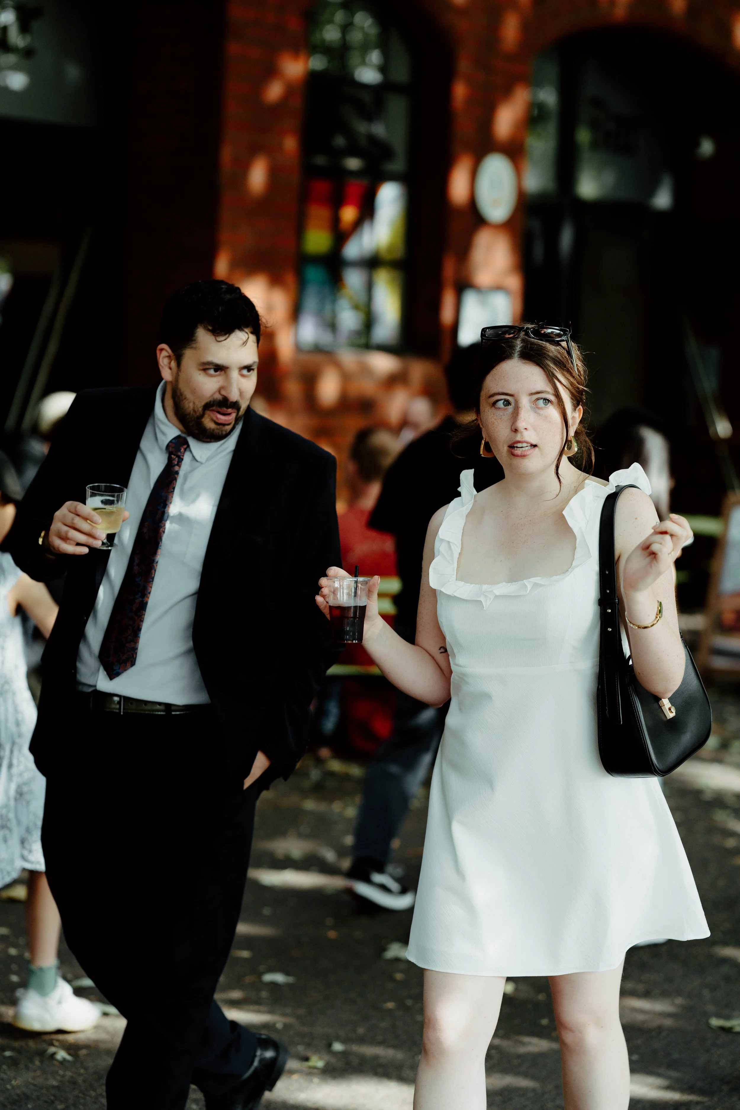 A man in a suit and a woman in a white dress are talking outdoors, holding drinks, with a brick building and other people in the background.