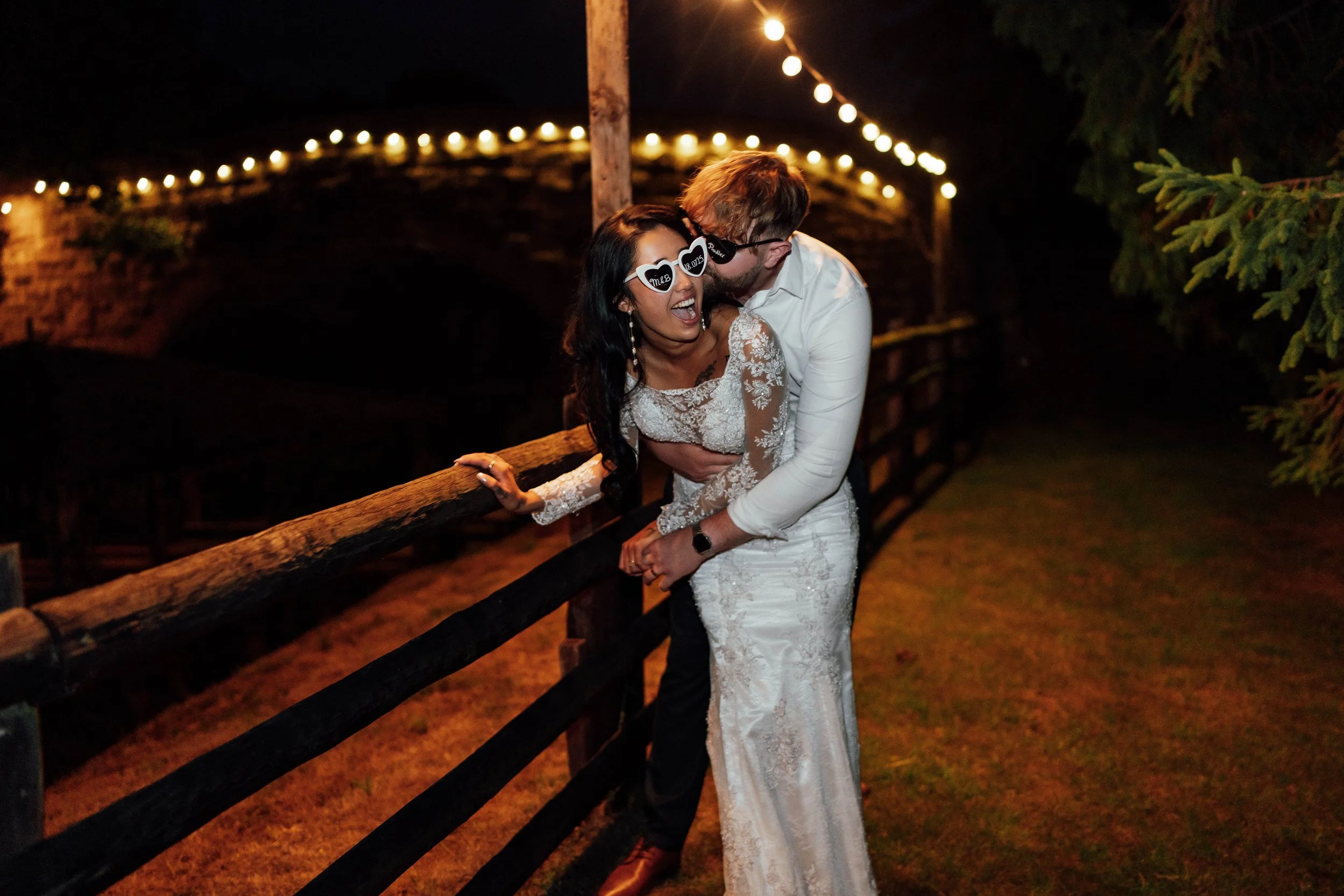 A joyful bride and groom in wedding attire enjoying themselves at night near a wooden fence, decorated with string lights overhead.