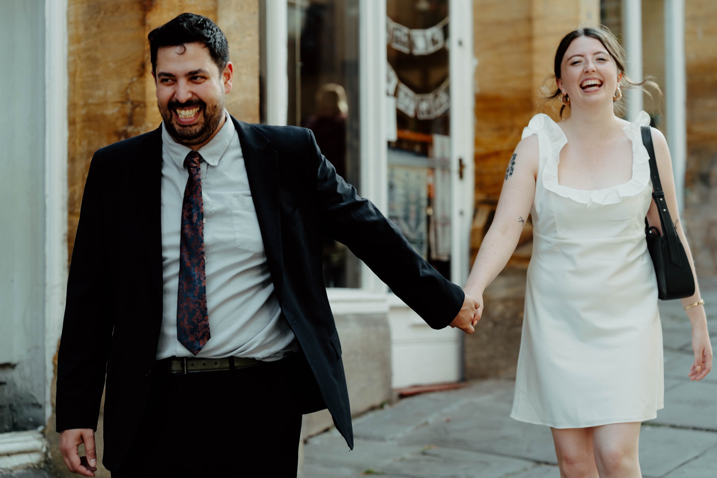 A man and a woman are holding hands and walking outside, both are smiling and appear happy. The man is dressed in a black suit with a white shirt and a patterned tie, while the woman is wearing a white dress with ruffled sleeves and carrying a black 