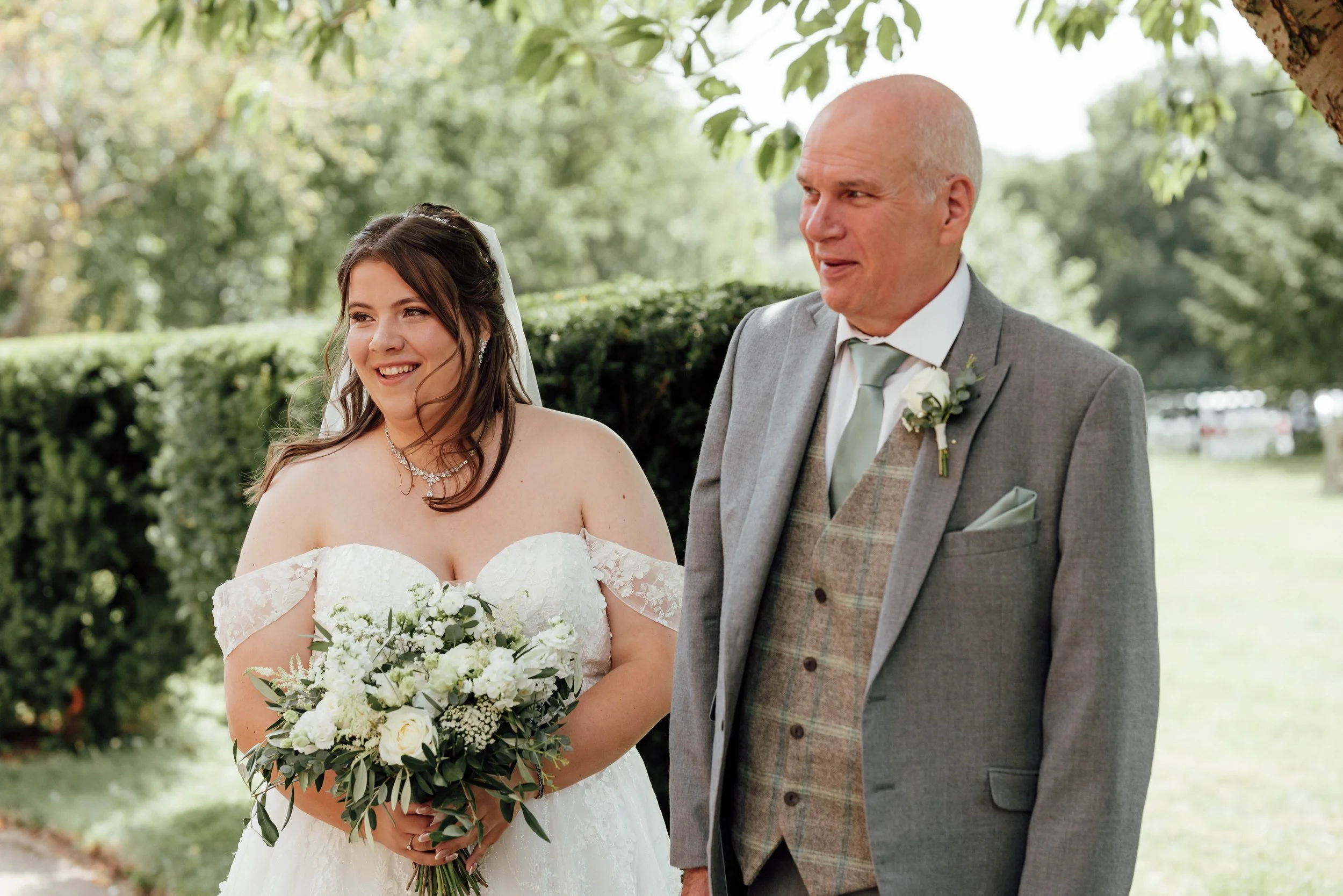 A bride holding a bouquet of white flowers stands beside an older man in a gray suit outdoors, with green trees and grass in the background.