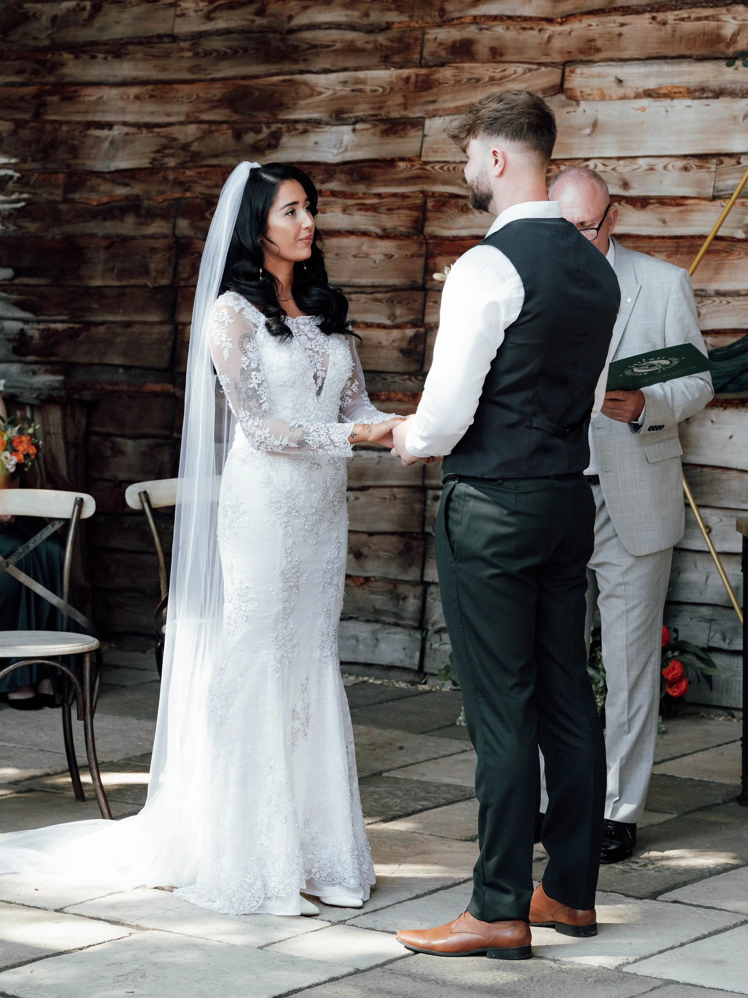 A bride and groom holding hands during their wedding ceremony, with an officiant reading from a book in a rustic setting with wooden walls.
