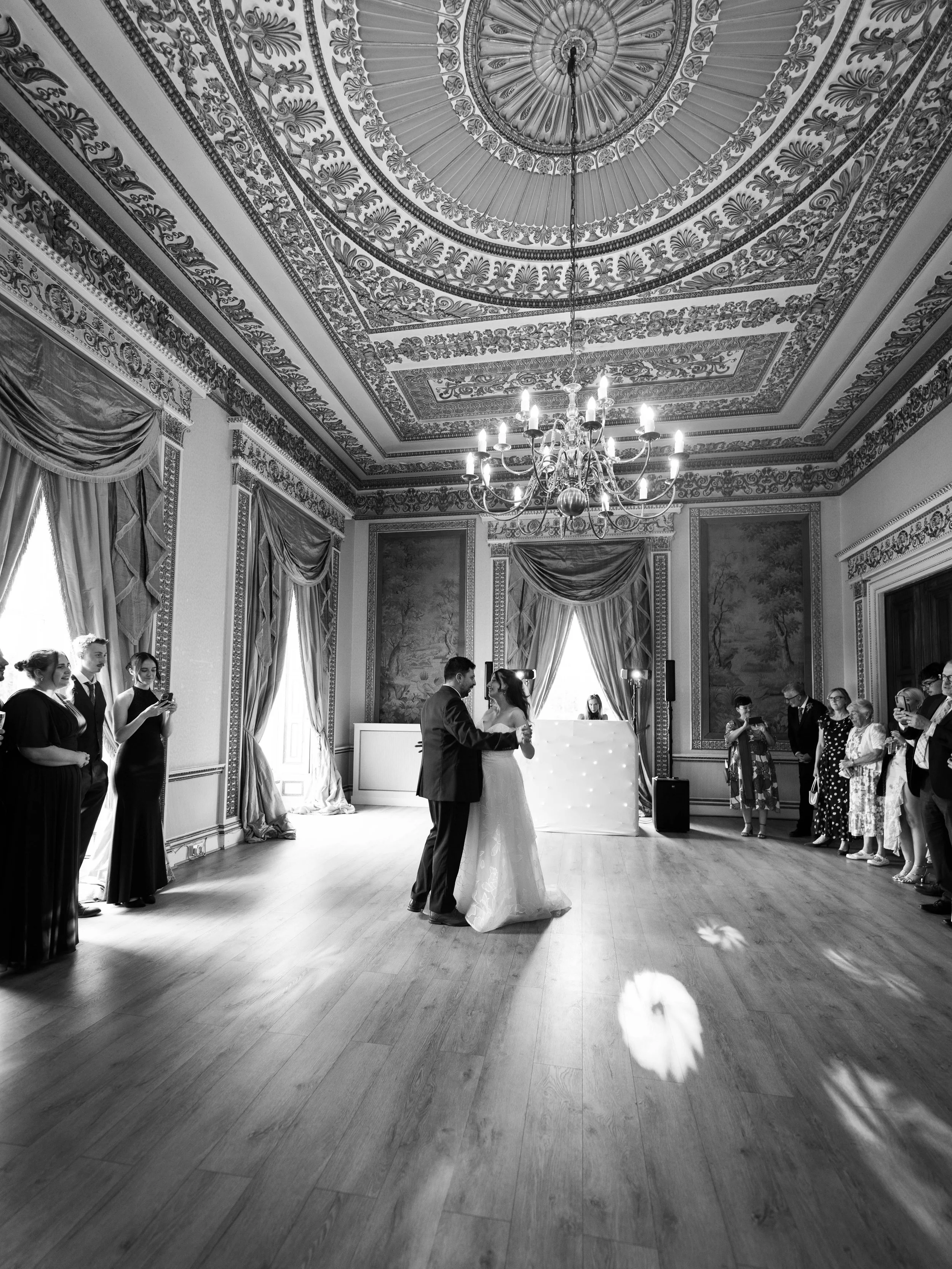 A black and white photo of a bride and groom dancing in a grand, ornate room with high decorated ceilings, large windows with draped curtains, and a chandelier. Guests are standing along the walls watching.