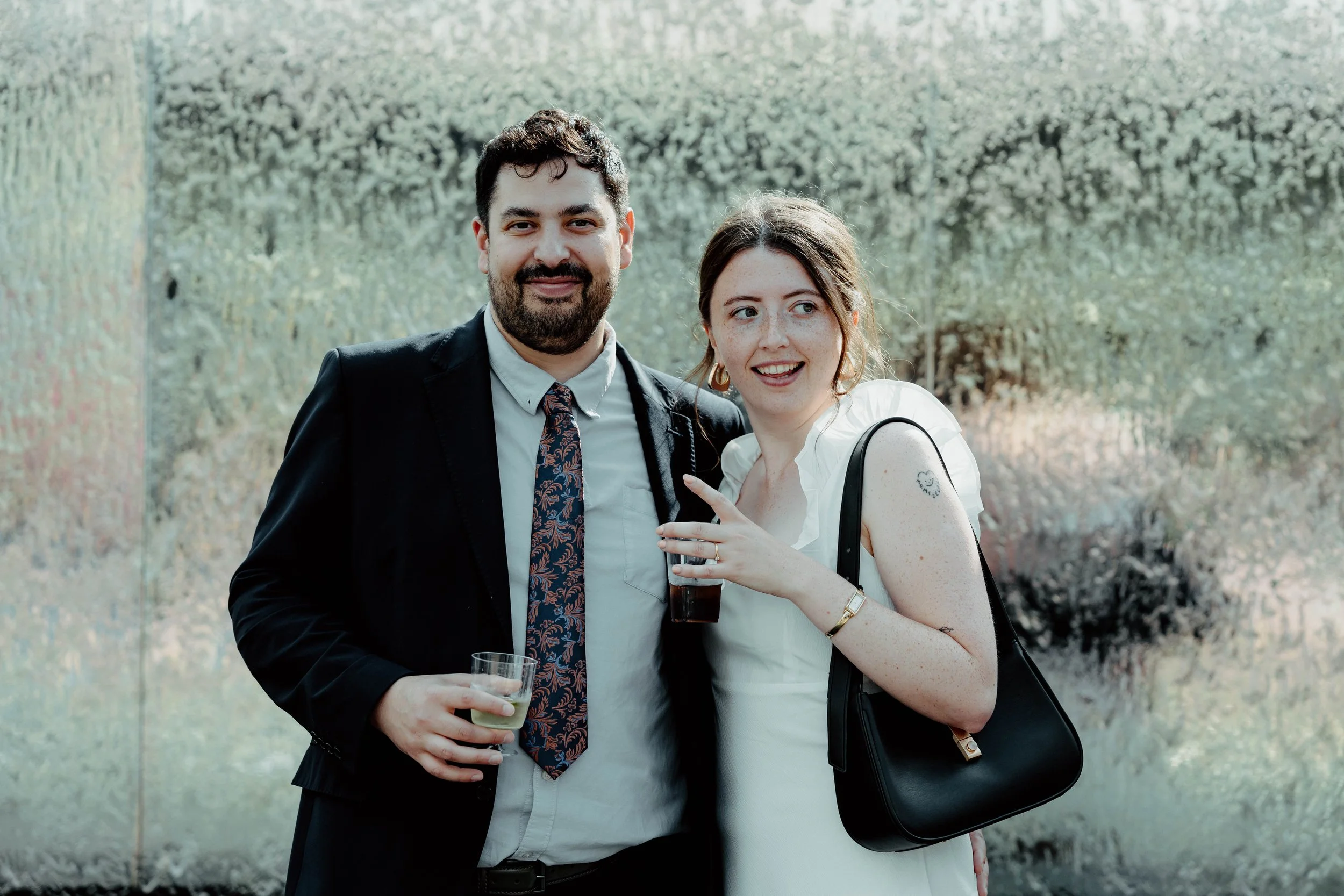 A man in a black suit holding a drink, and a woman in a white dress holding a drink, standing outdoors in front of greenery, smiling and enjoying a social event.